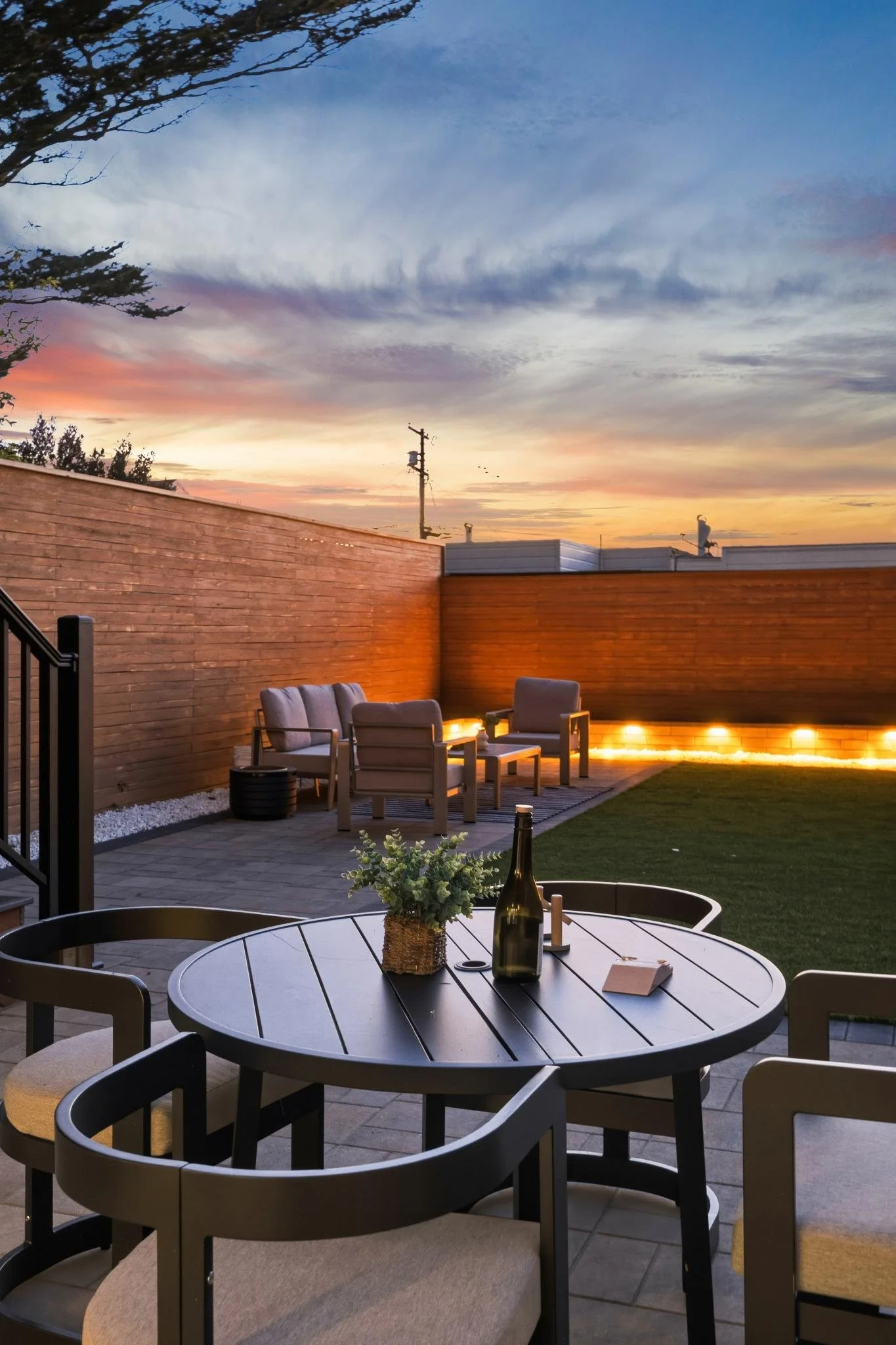 Outdoor patio with a round dining table, chairs, a bottle of wine, and a small plant, with a seating area in the background under a colorful sunset sky.
