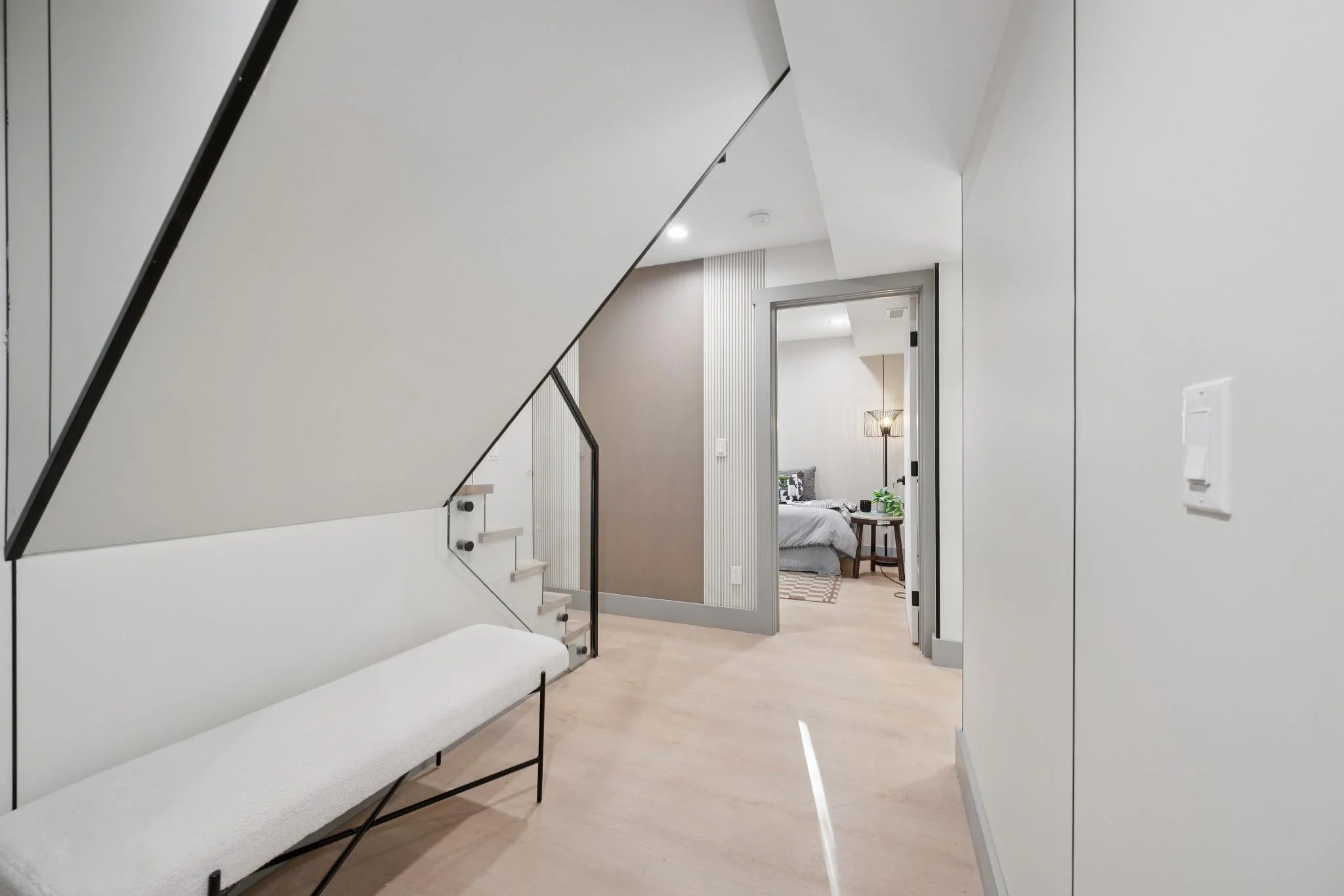 Interior view of a modern home with a hallway leading to a bedroom. The hallway features a bench with a white cushion, a staircase with white steps and black railing, and beige flooring. The bedroom is furnished with a bed, a nightstand, and a standi