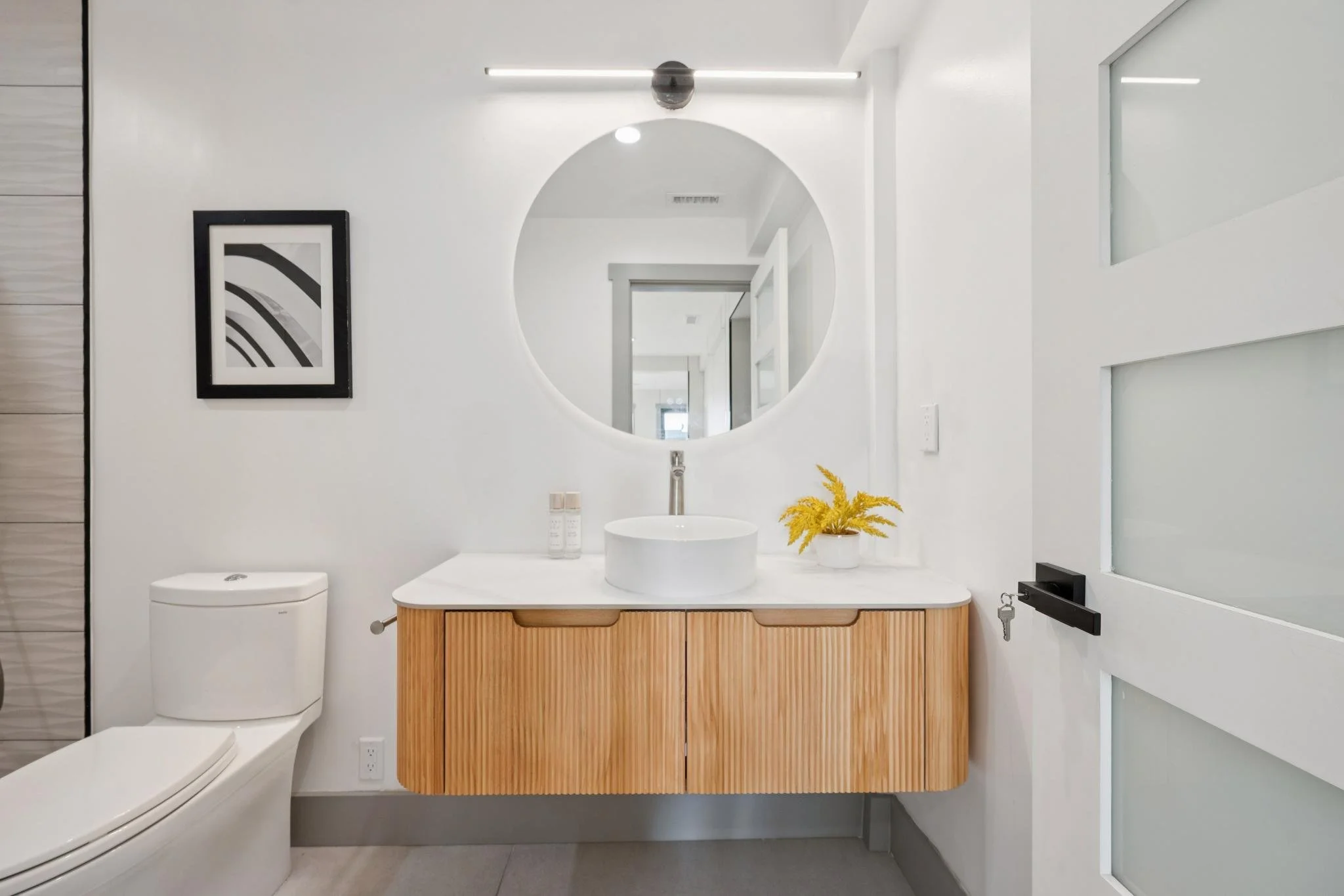 Modern bathroom with a white floating vanity, a round mirror, a vessel sink, and a decorative yellow plant. To the left, a white toilet, and to the right, a slide door with frosted glass panels.