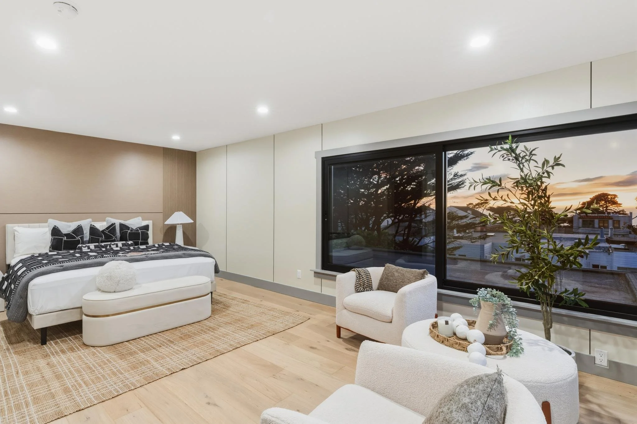 A modern bedroom with a white bed, black and white pillows, a beige bench, a beige armchair with a brown pillow, a wicker tray with white balls and a plant on a white round table, a large window with trees outside at sunset, and warm wooden flooring.