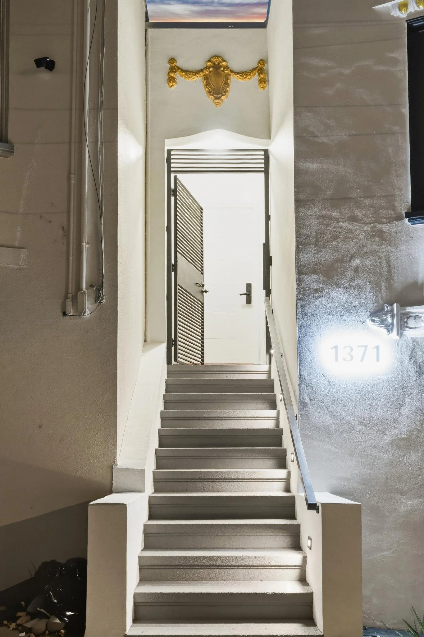 Indoor staircase leading to a door with windows at the top, all painted white. A gold decorative crest is above the door, and the number 1371 is visible on the wall to the right.