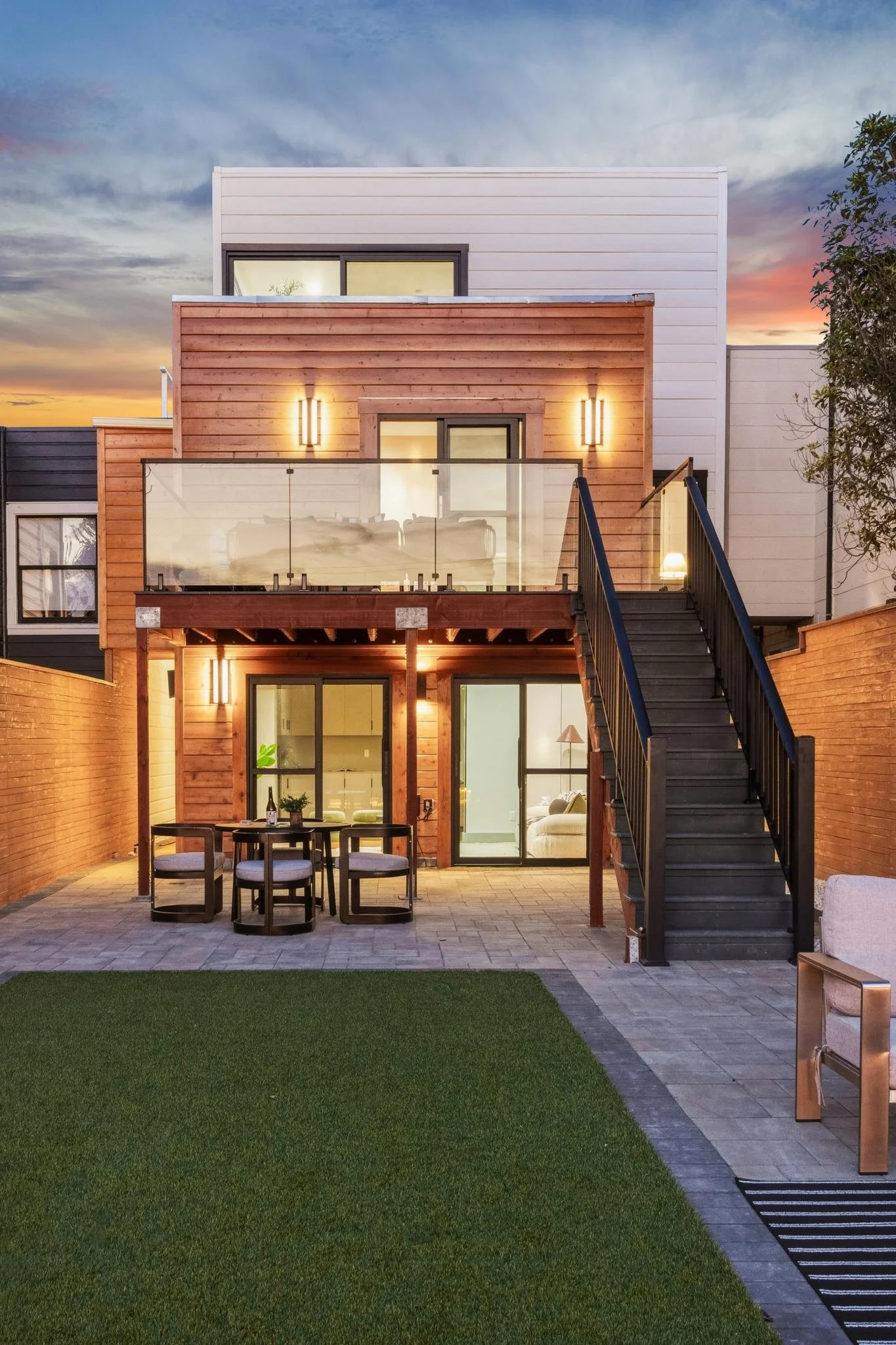 Modern multi-story house with wooden and white exterior, illuminated outdoor lighting, glass balcony railing, staircase leading to upper deck, patio with dining table and chairs, and a lawn area at sunset.