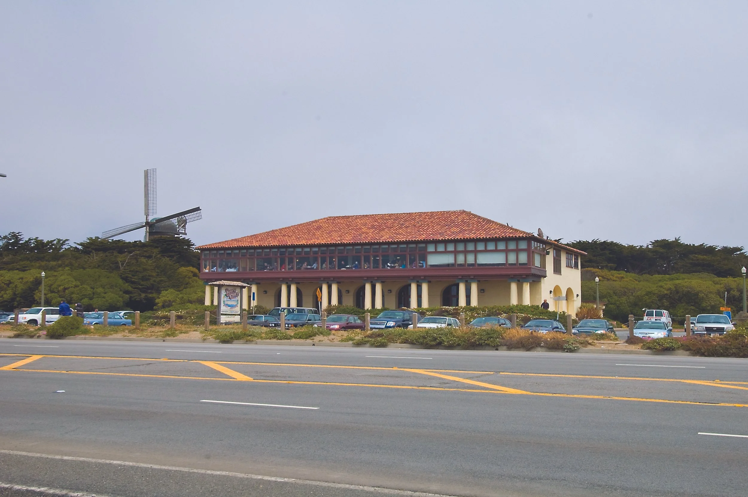 A white two-story building with a red-tiled roof and a large wraparound windowed upper floor, situated near a coastal road with multiple parked cars and a windmill in the background.