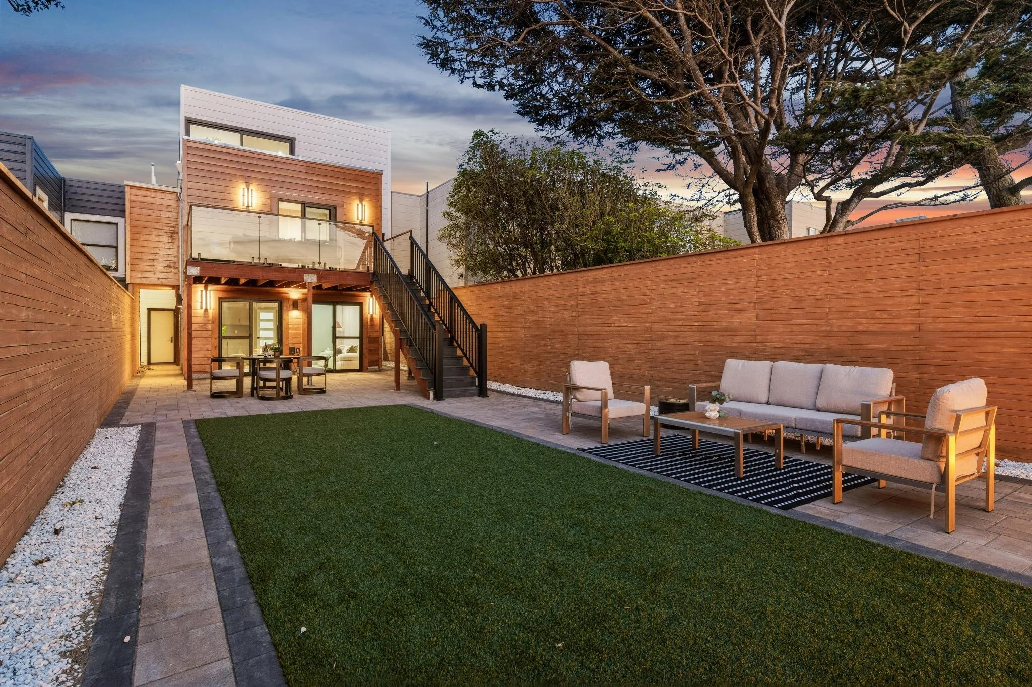 A modern backyard patio at dusk with outdoor furniture including a sofa and chairs on a rug, a small table, a dining table with chairs, and a attached two-story house with a staircase leading to a upper deck, all enclosed by a tall wooden fence and s