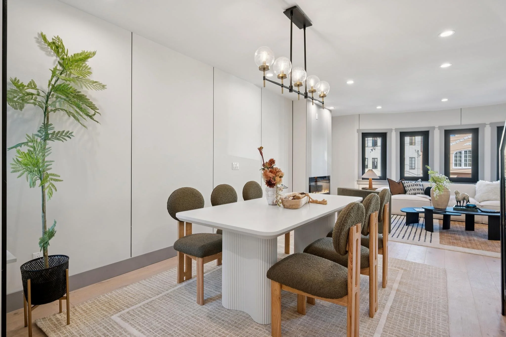 Modern dining and living room with white walls, wooden flooring, a white dining table with six brown chairs, a black and gold chandelier, and a living area with a white sofa, a black coffee table, and large windows.