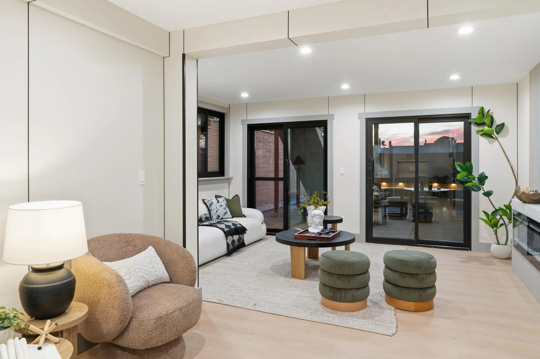 Living room with white walls, hardwood flooring, and black-framed sliding glass doors leading to an outdoor area. Contains a white sofa with pillows, a brown rounded armchair with a pillow, a black round coffee table, two green round ottomans, a pott