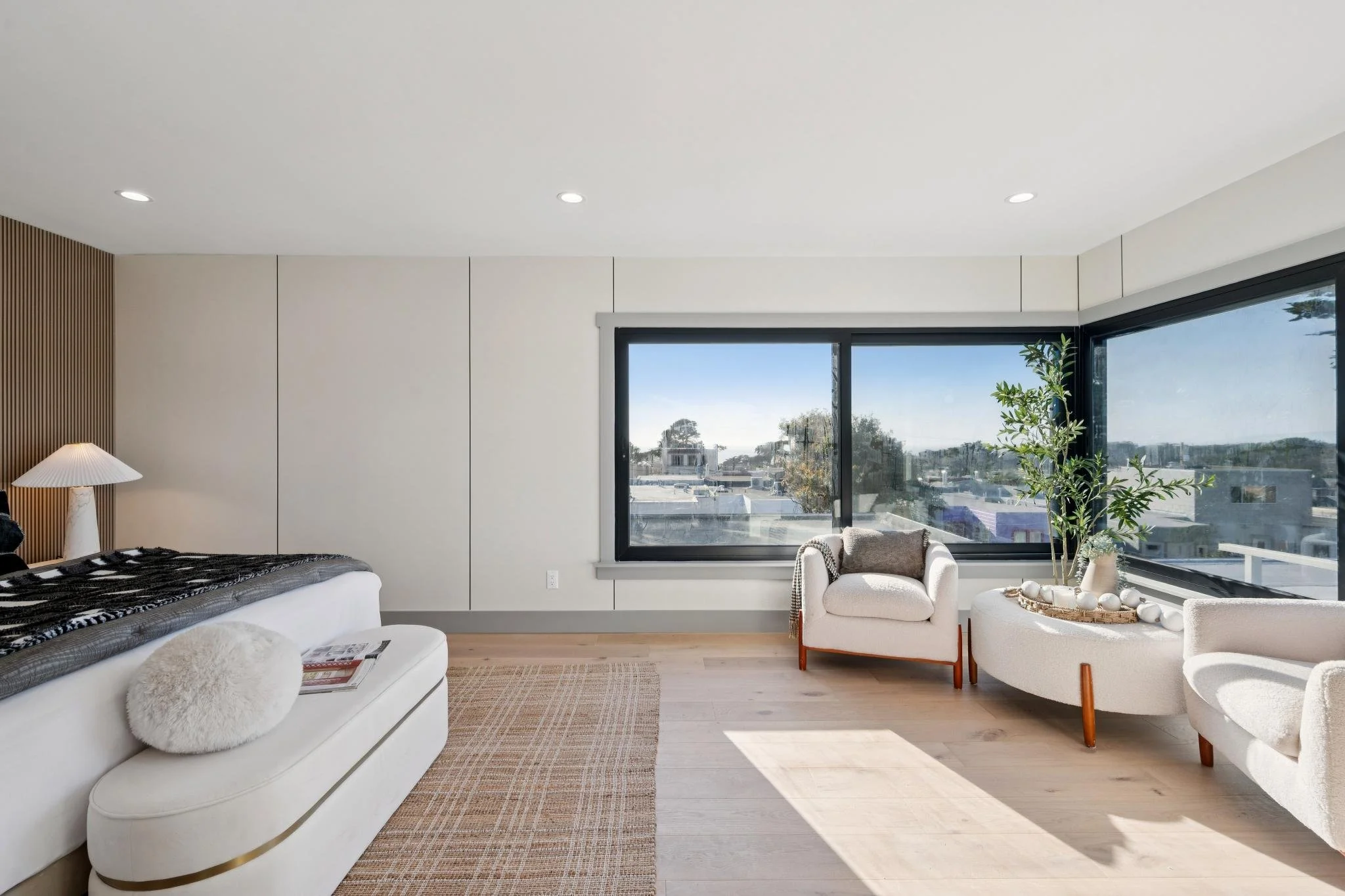 Bright bedroom with large windows, a bed with black and white bedding, a white bench with a pillow, armchairs, a tall plant, and natural light.
