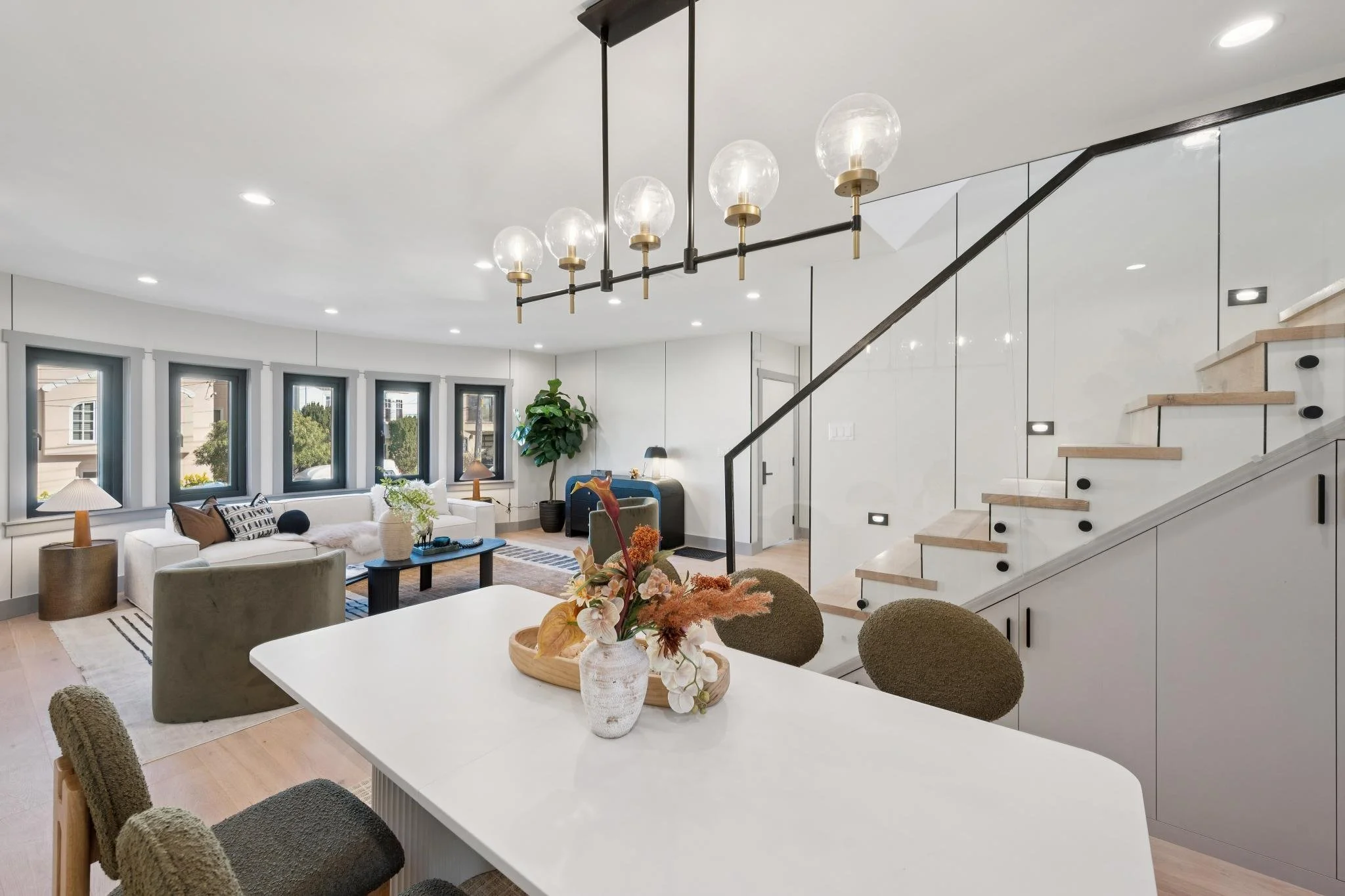 Open-concept living room and dining area with modern decor, multiple windows, a staircase with a black railing, a large white table with a flower arrangement, and a cozy seating area with sofas and lamps.