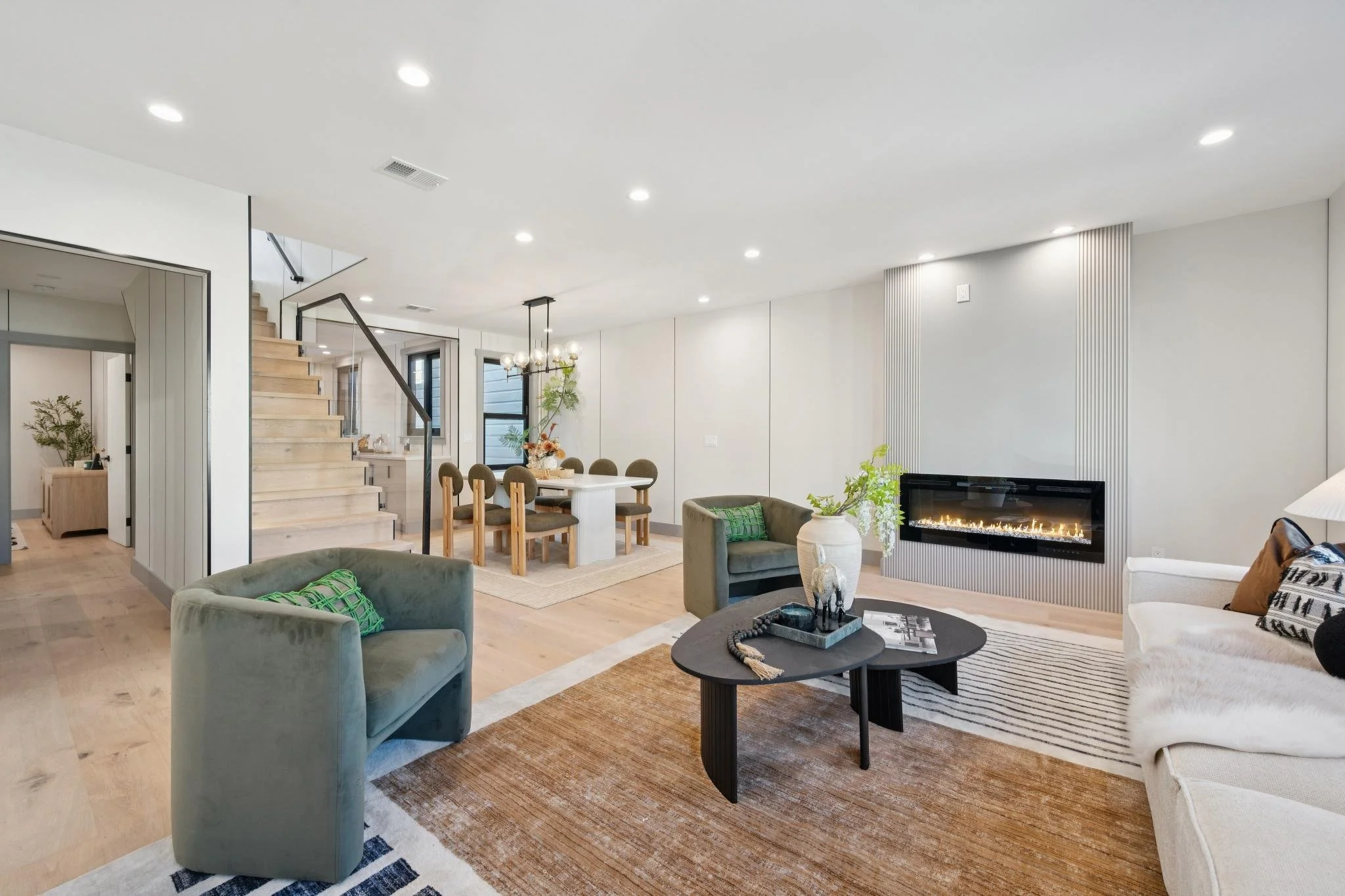Modern living room with a beige sofa, green armchairs, black coffee table, fireplace, and dining area with wooden table and chairs.
