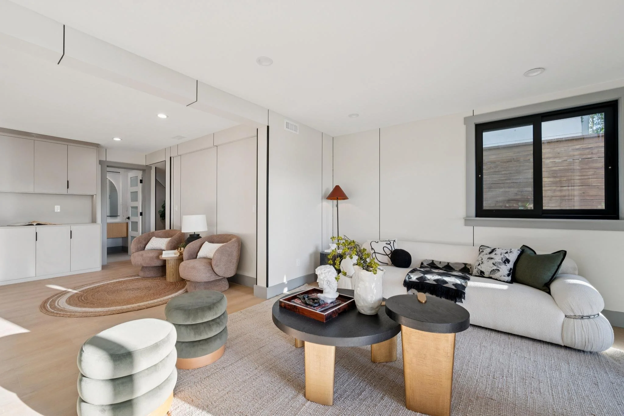 Bright living room with beige walls, a large white sofa with black and white pillows, a dark wood oval coffee table with vases and decorative items, two light pink chairs with cushions, a rattan side table, a green plush ottoman, a window overlooking