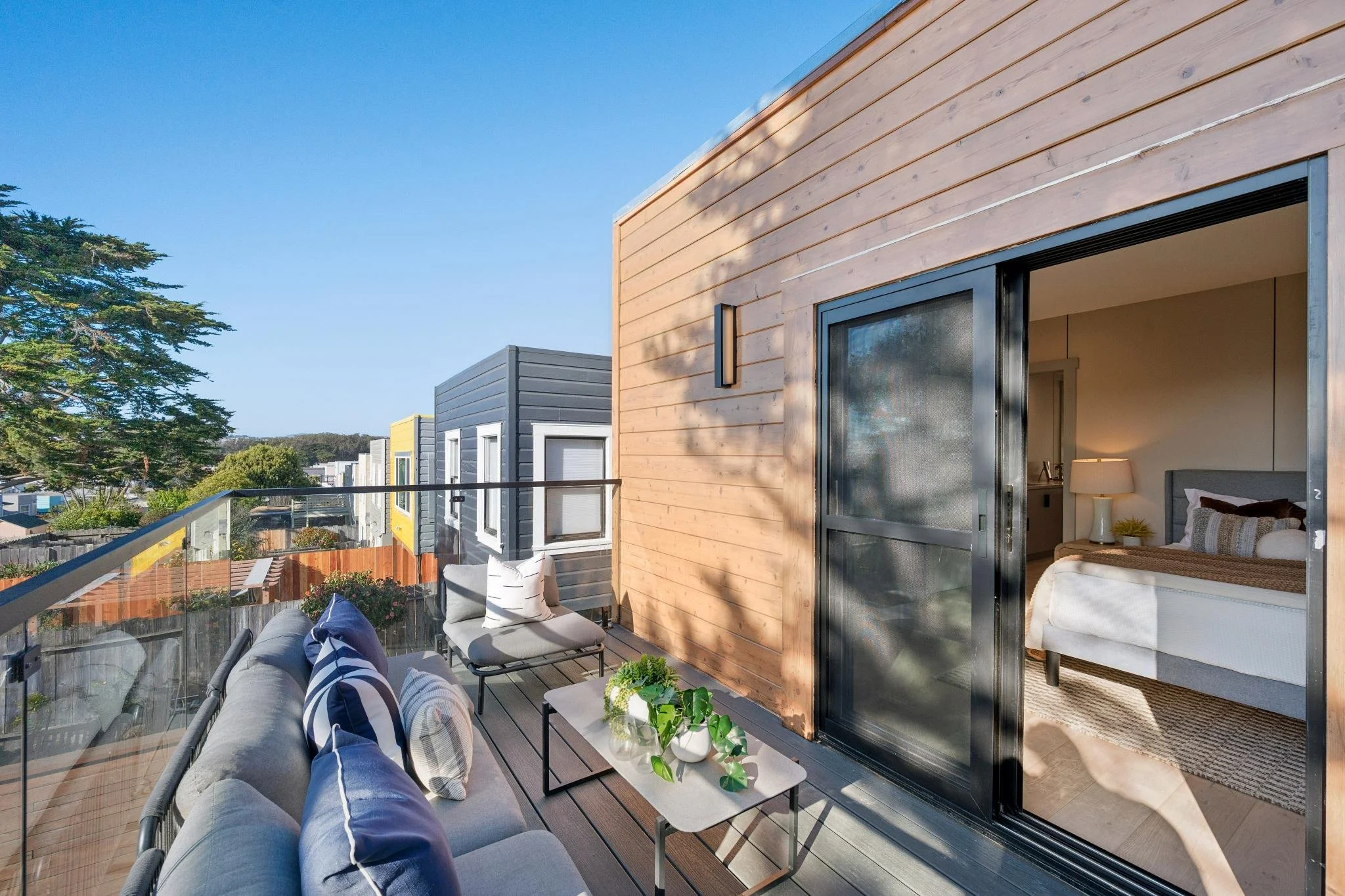 Balcony of a modern house with outdoor seating, pillows, and a small table with plants. Open sliding door to a bedroom with a bed, nightstand, and lamp.