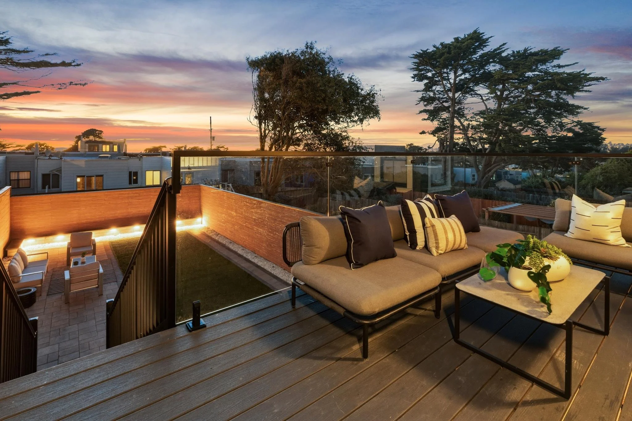 Outdoor rooftop patio at sunset with cushioned seating, a coffee table with potted plants, and a view of trees and the colorful sky.