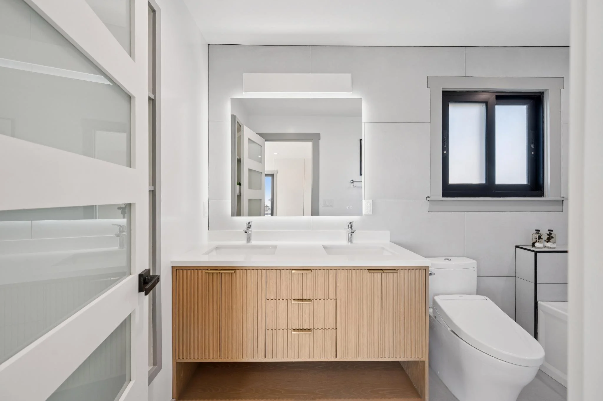Modern bathroom with double vanity, frameless mirror, black window, and toilet.