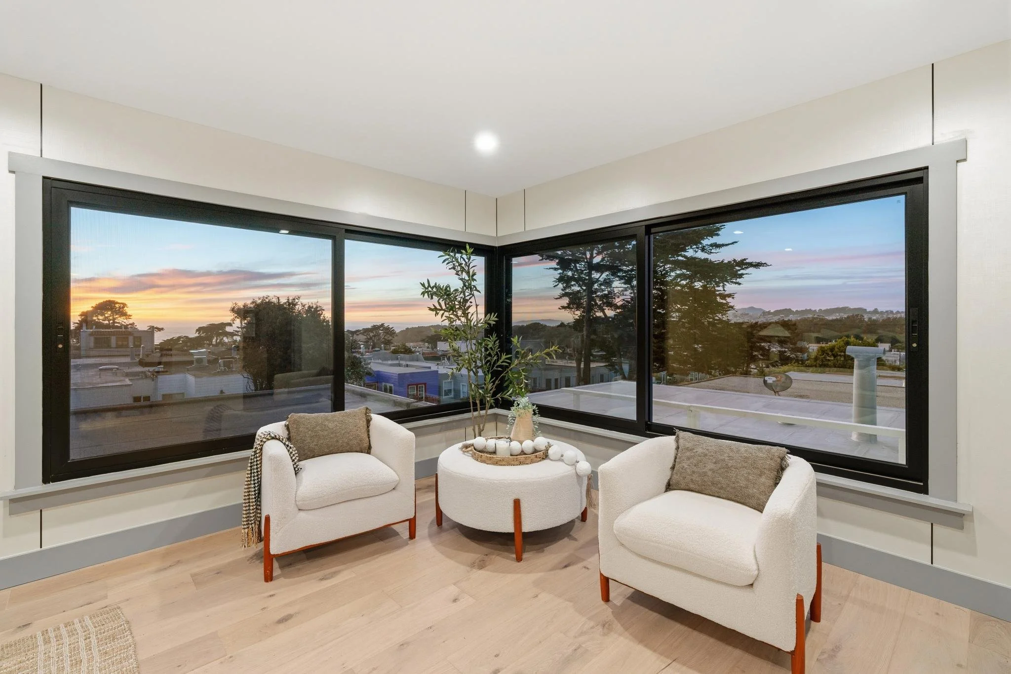 Interior of a room with two white armchairs, a round coffee table with decorative plants and spheres, large corner windows showing a sunset over rooftops and trees.