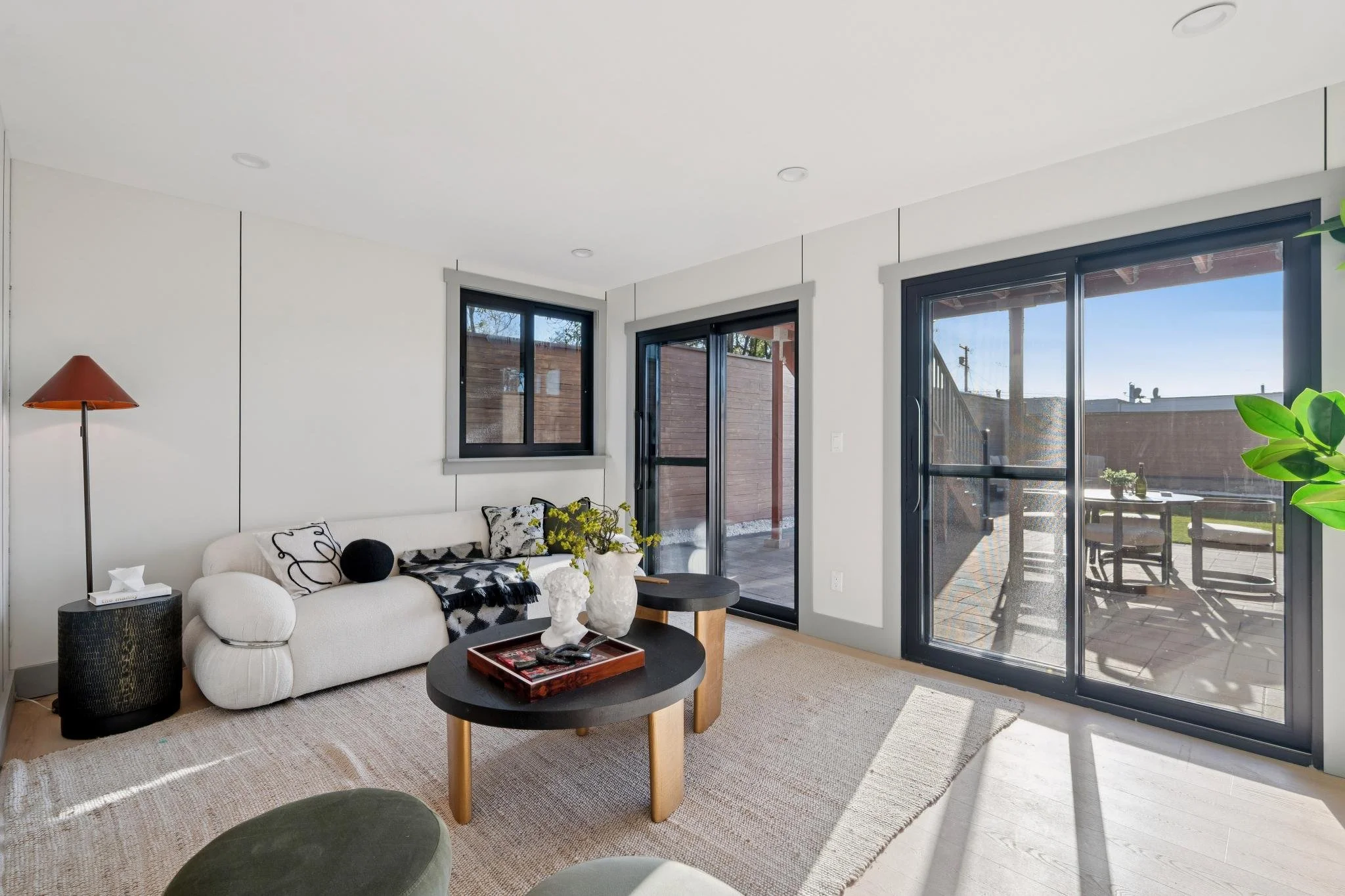 Living room with white sofa, black and white pillows, black round side table, coffee table with decorative items, large sliding glass doors opening to patio, window panes, and a rug on light-colored wood floor.