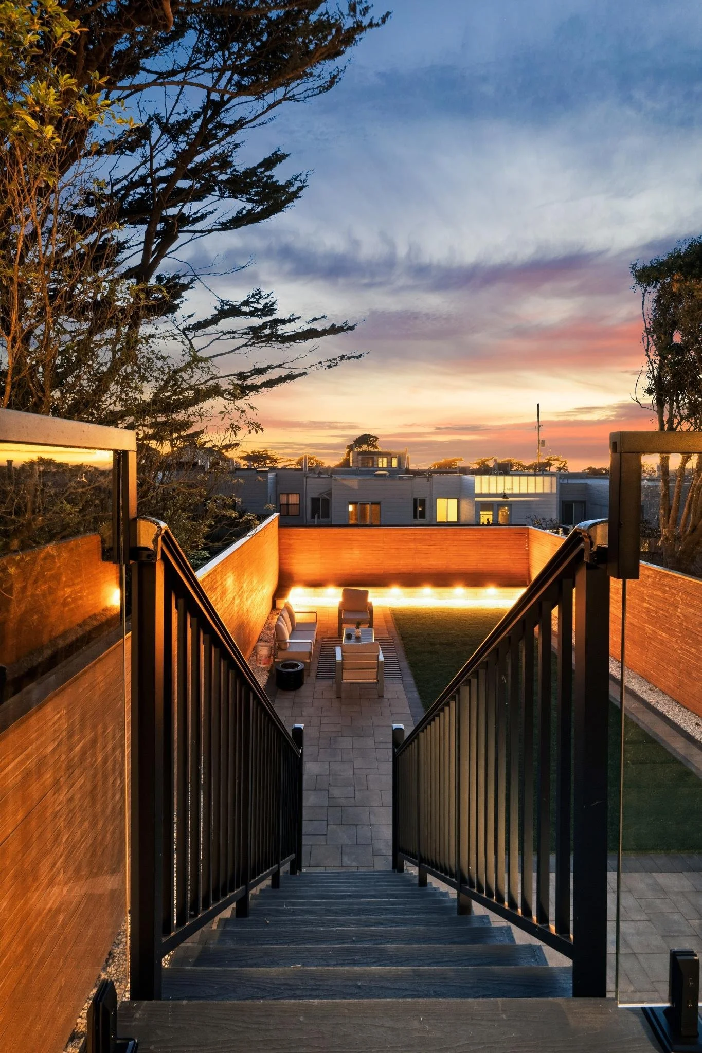 A view from the top of a staircase overlooking a backyard patio with outdoor furniture, a brick wall, and a sunset sky.