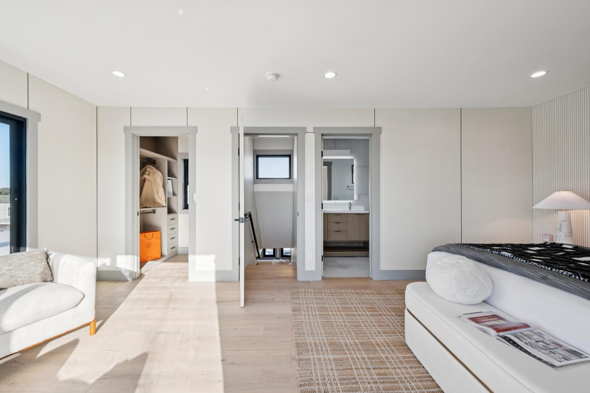 Modern bedroom with white walls, light wood flooring, a bright window, a white sofa, a bed with a pillow, and a lamp, with doorways leading to a closet, staircase, and bathroom.
