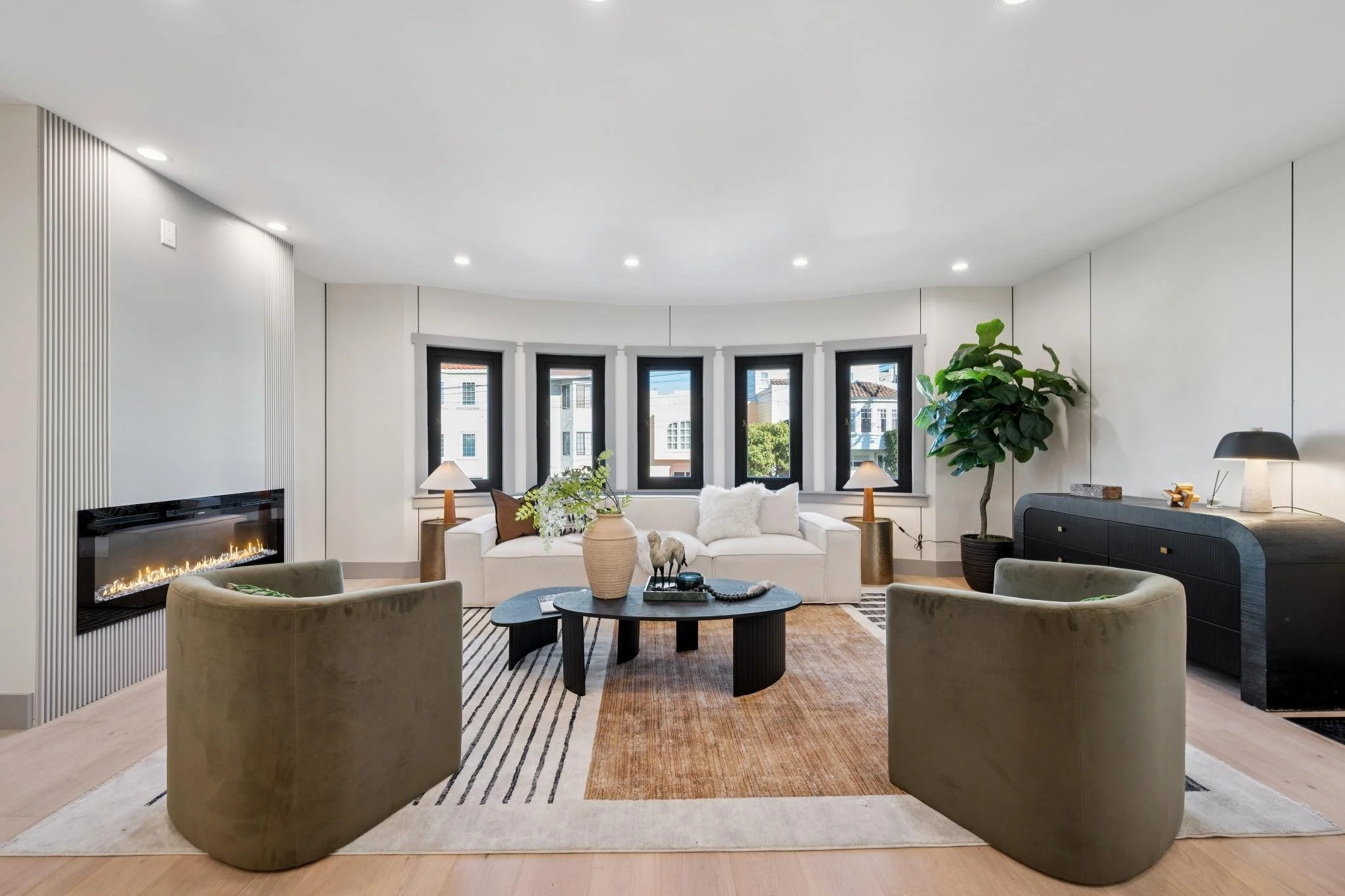 Modern living room with a white sofa, two green armchairs, a black coffee table, a fireplace, and large bay windows with black frames, decorated with lamps, plants, and decorative items.