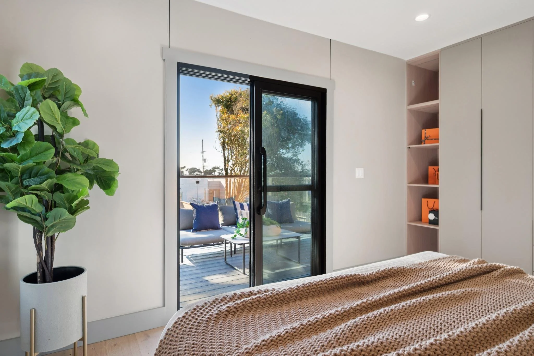 A bedroom with a sliding glass door leading to an outdoor patio with a seating area. The bedroom has a bed with a textured beige blanket, a potted green plant on the left, and built-in shelving with orange boxes on the right.
