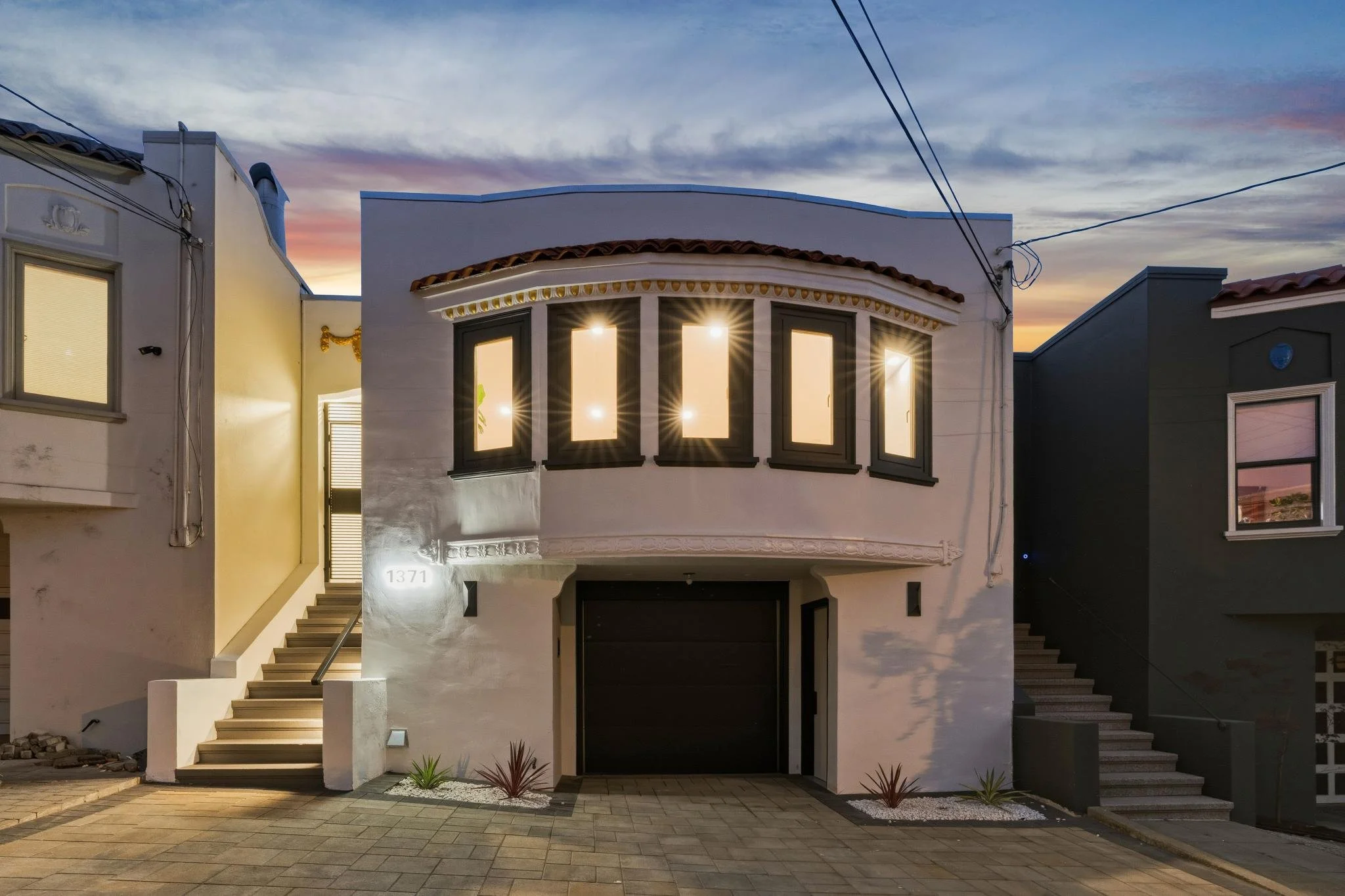 A modern white two-story house with a curved upper facade and large windows, illuminated from inside, located between two other houses during twilight.