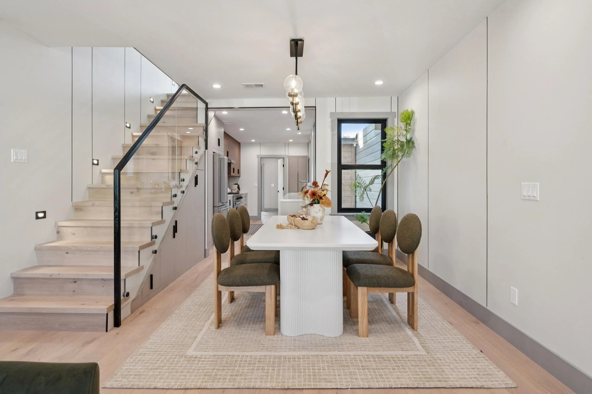 Modern dining room with a white table, eight green upholstered chairs, a light wood floor, and a staircase on the left side. A large window with black trim is at the back, with plants on the right side.
