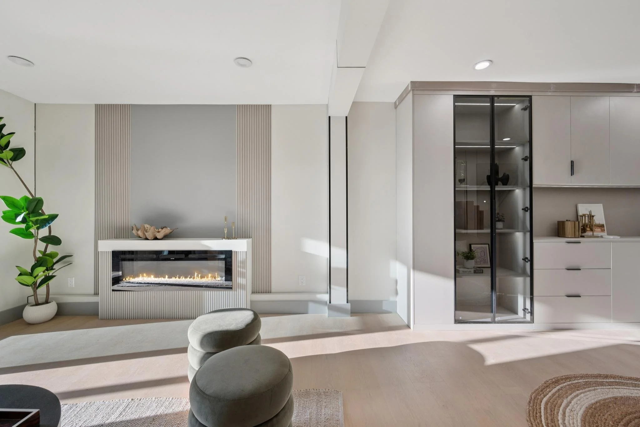 Modern living room with electric fireplace, potted plant, white furniture, glass cabinet, and wooden accents, illuminated by natural light.