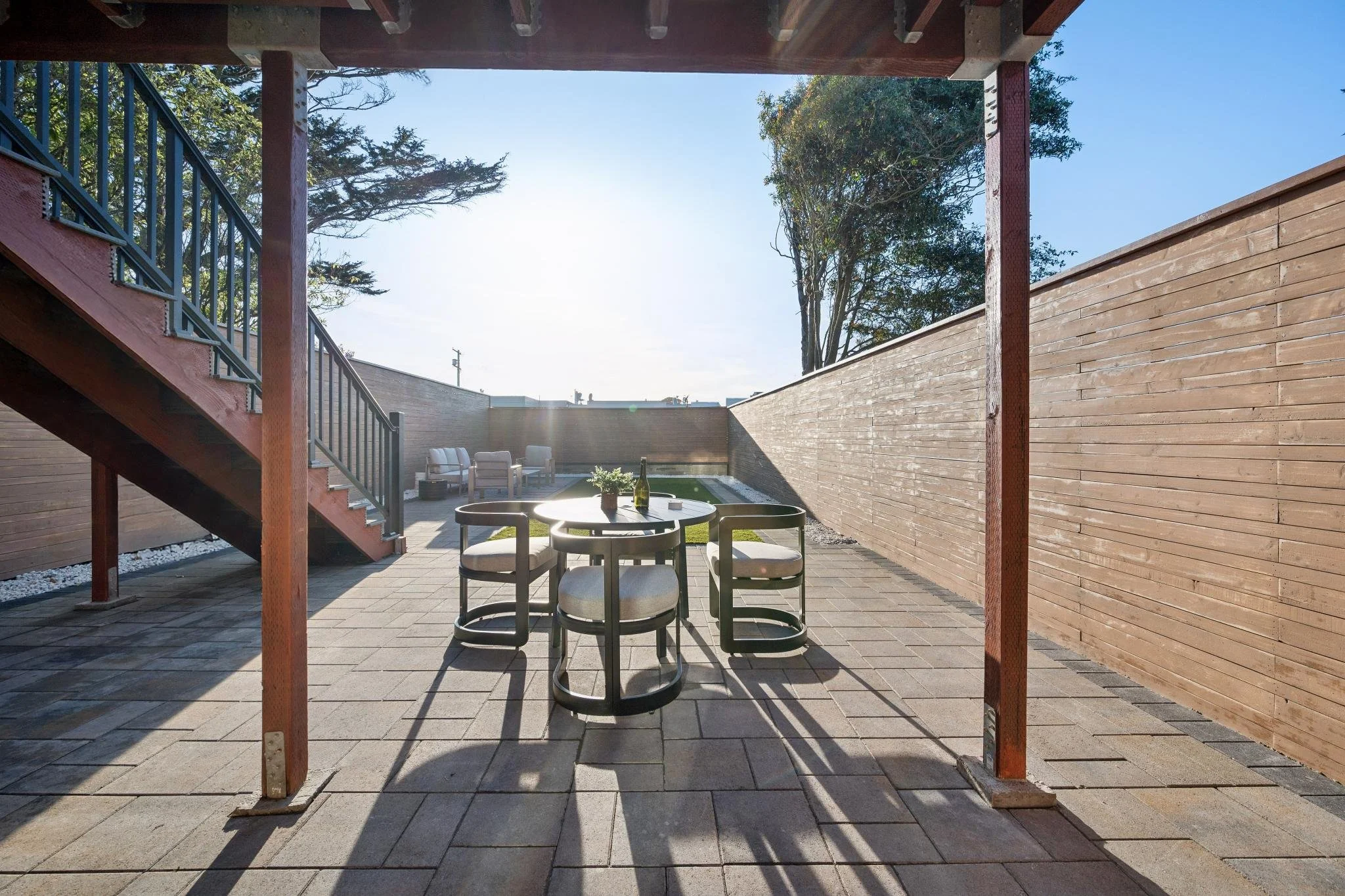 Back patio with a round table and four chairs, a small swimming pool, a couple of lounge chairs, wooden fencing, and a staircase leading to an upper level, with trees in the background.