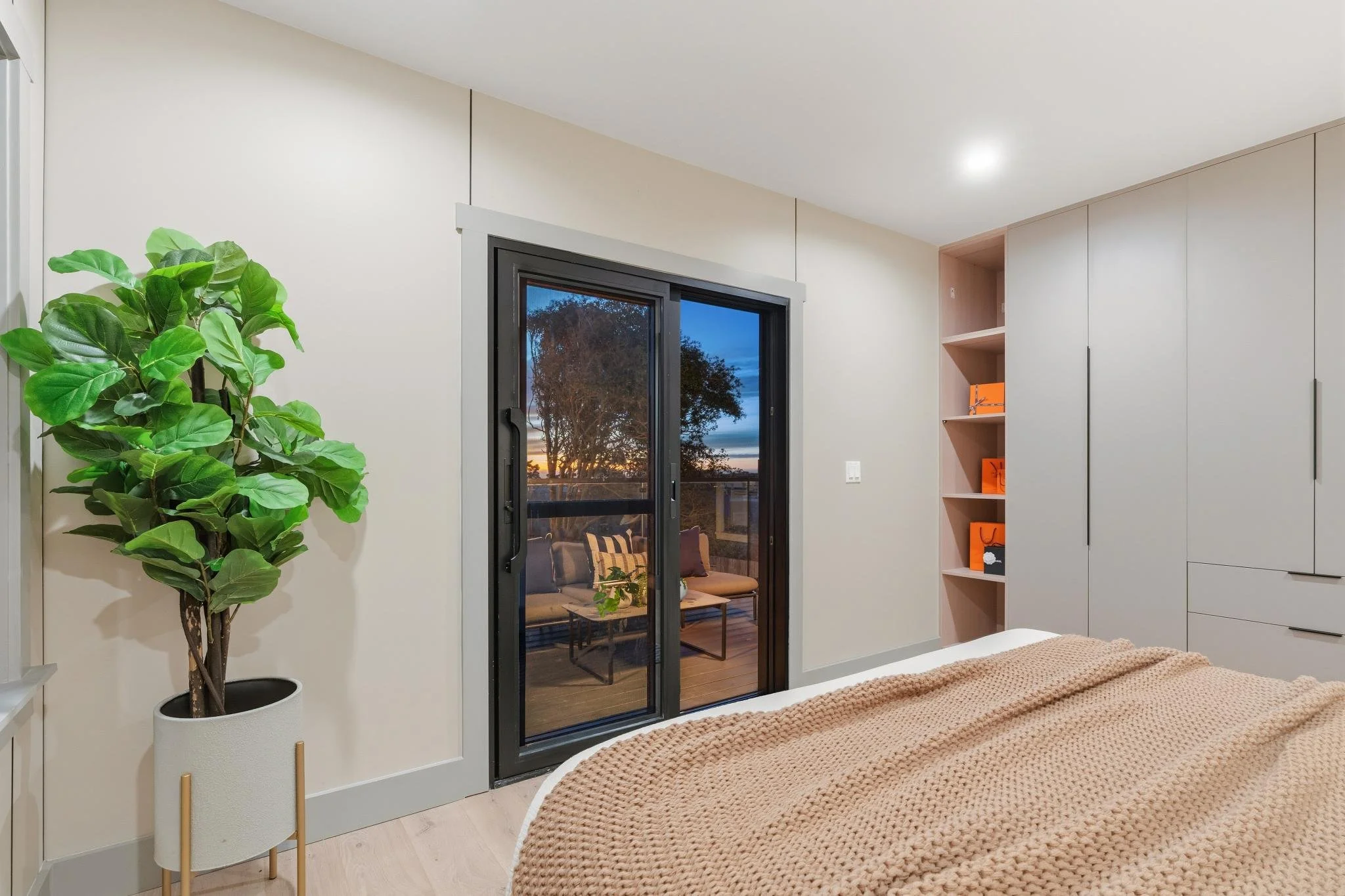 A bedroom with a large potted fiddle leaf fig plant, a sliding door to a balcony, a knitted blanket on the bed, and built-in white closets with orange decor accessories.