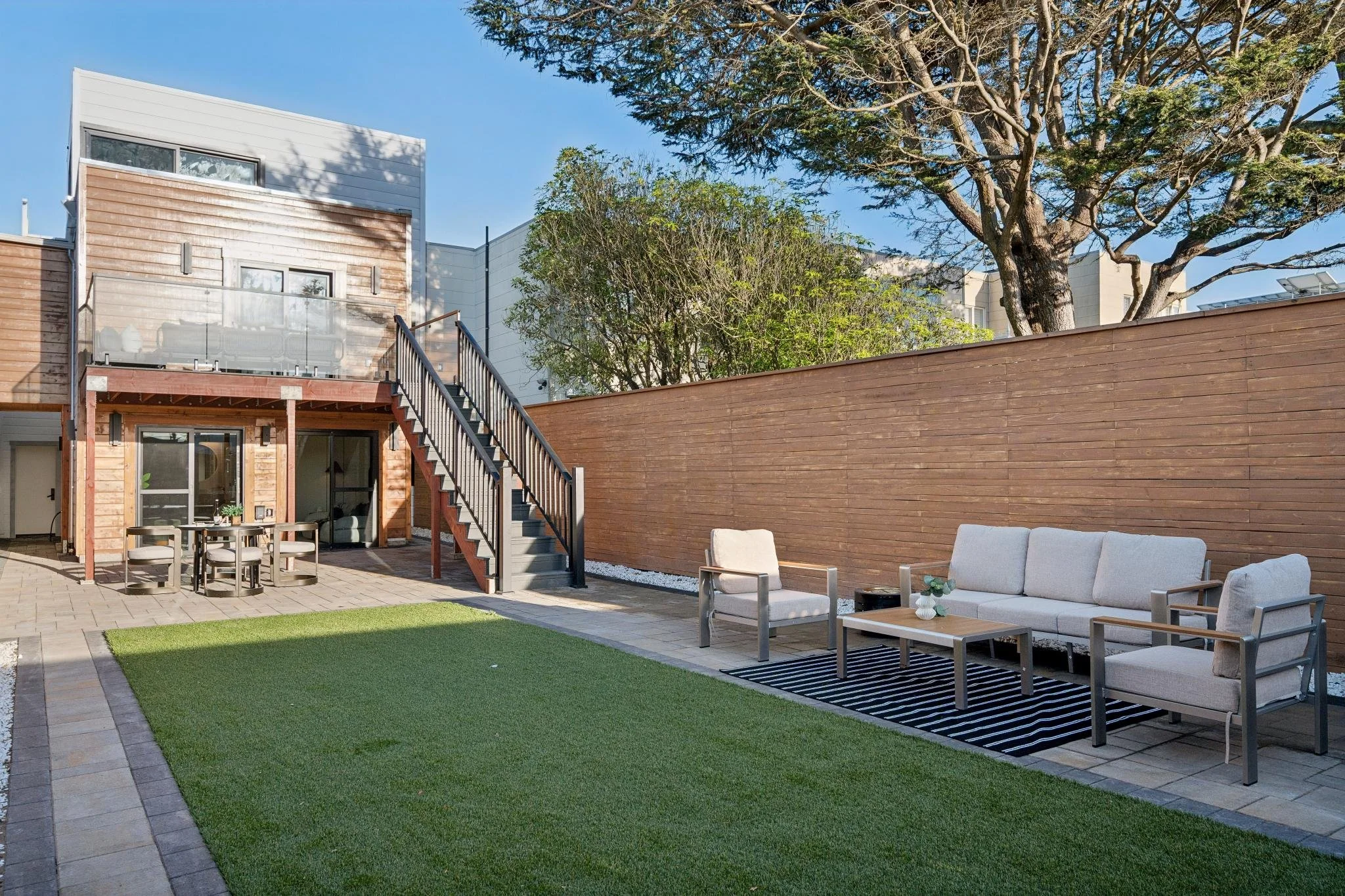 Clean outdoor backyard patio with seating area, outdoor dining table, staircase leading to upper deck, and tall wooden privacy fence, with trees and residential buildings in the background under clear blue sky.