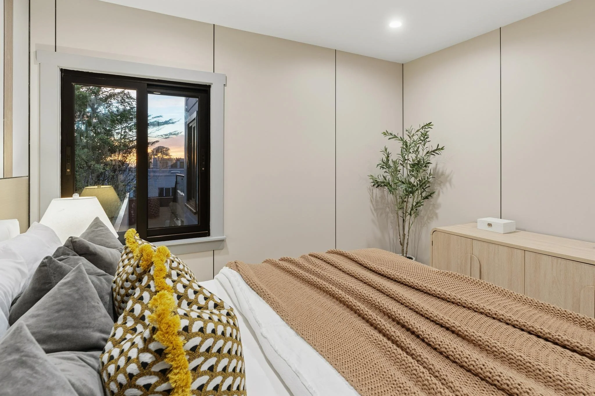 A neatly made bed with white and gray pillows, beige blanket, and a patterned pillow with yellow pom-pom detail. There is a window showing a sunset and a bedroom with minimal decor, including a light wood dresser and a potted plant.