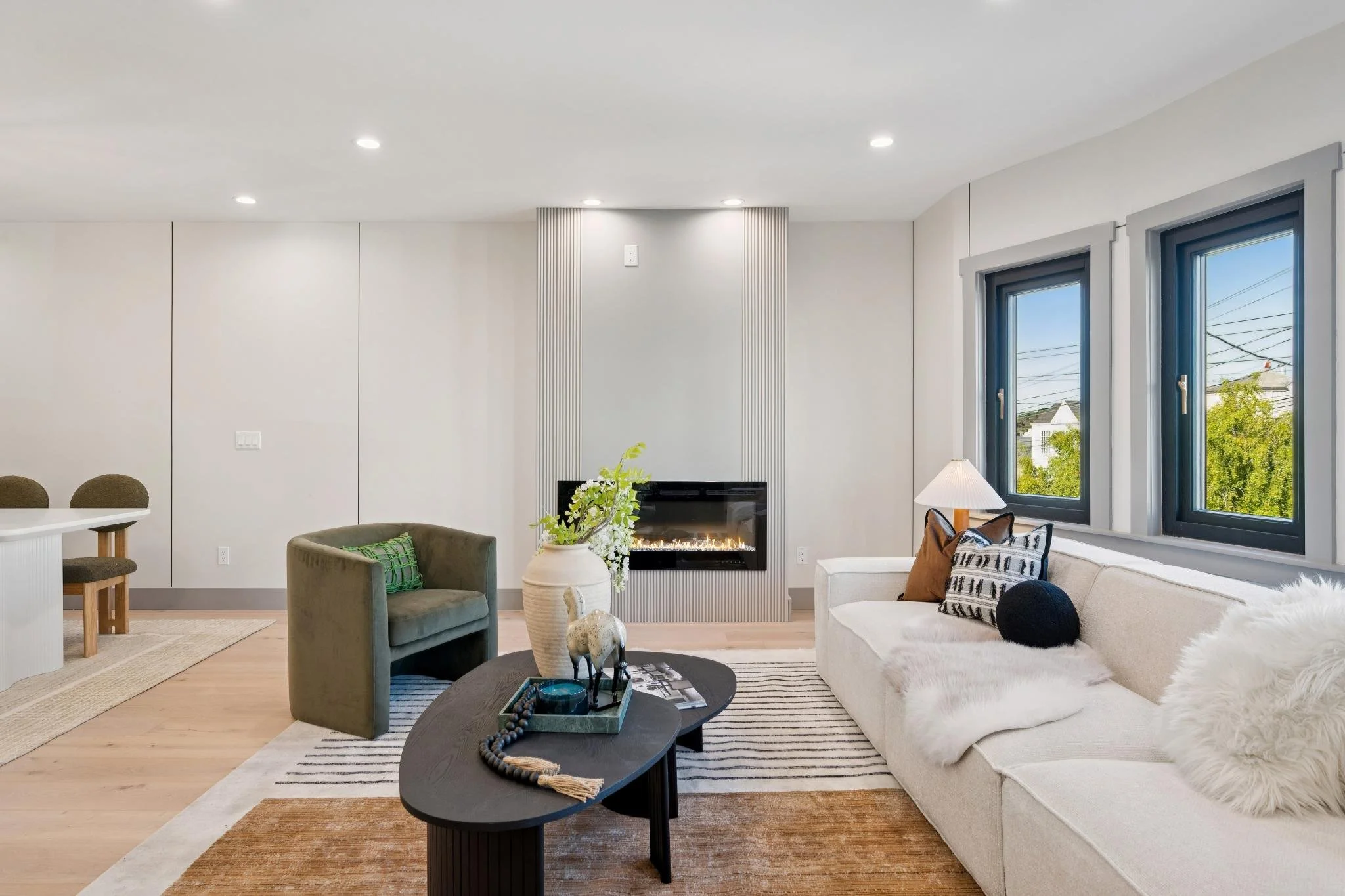 Living room with white walls, a white sectional sofa with decorative pillows and a throw blanket, a black coffee table with decorative items including a horse sculpture and plants, a green armchair with a pillow, a fireplace with vertical paneling, t