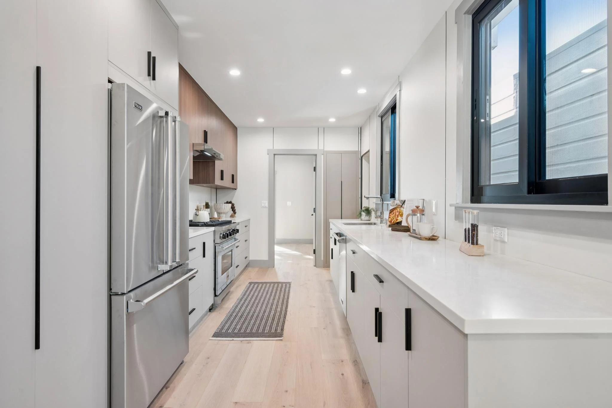 Modern kitchen with white cabinets, black handles, stainless steel appliances, and light wood flooring. Large windows on the right side let in natural light.
