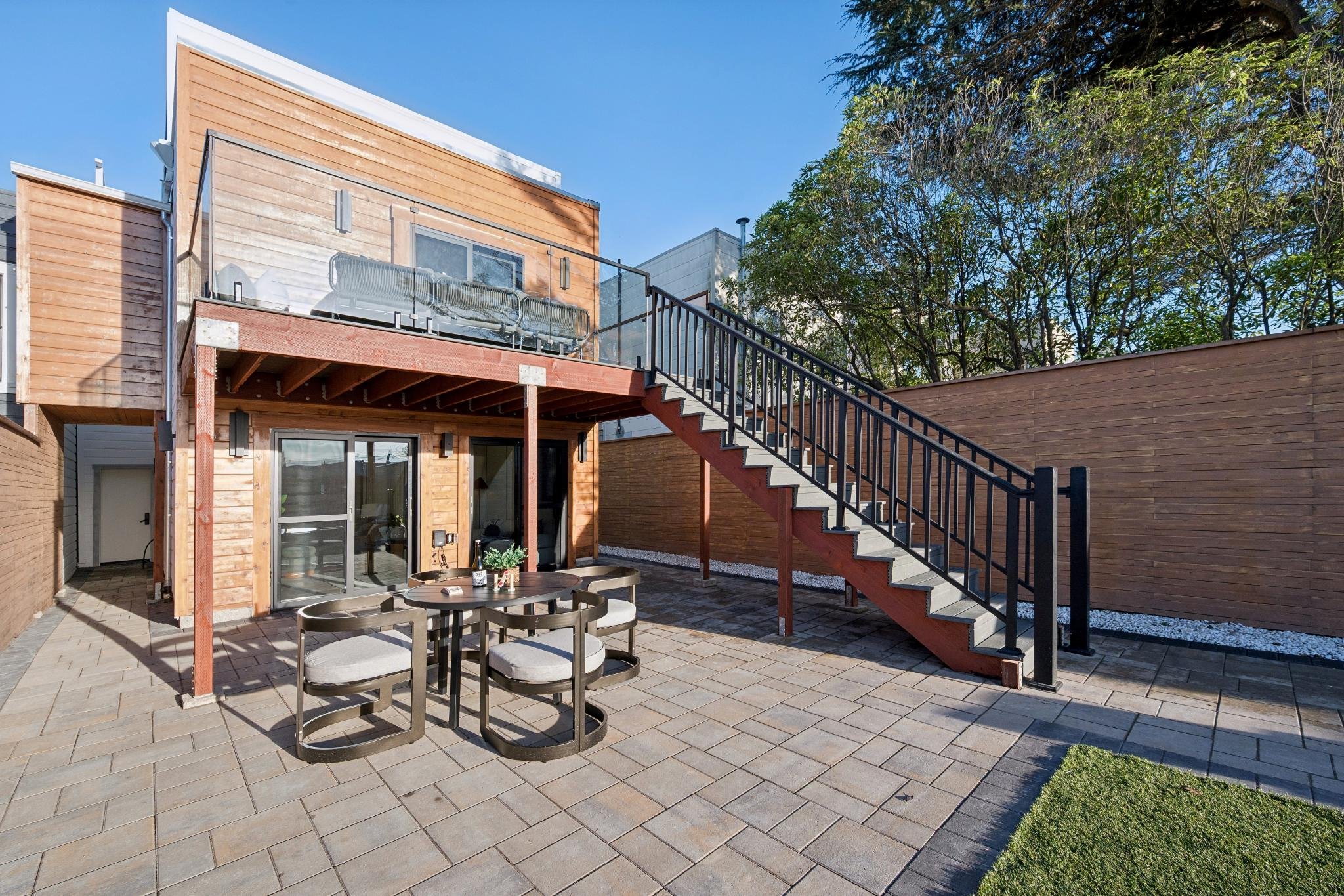 Backyard patio with outdoor dining table and chairs, wooden deck with glass railing, and stairs leading to upper level. Fenced yard with trees and shrubbery.