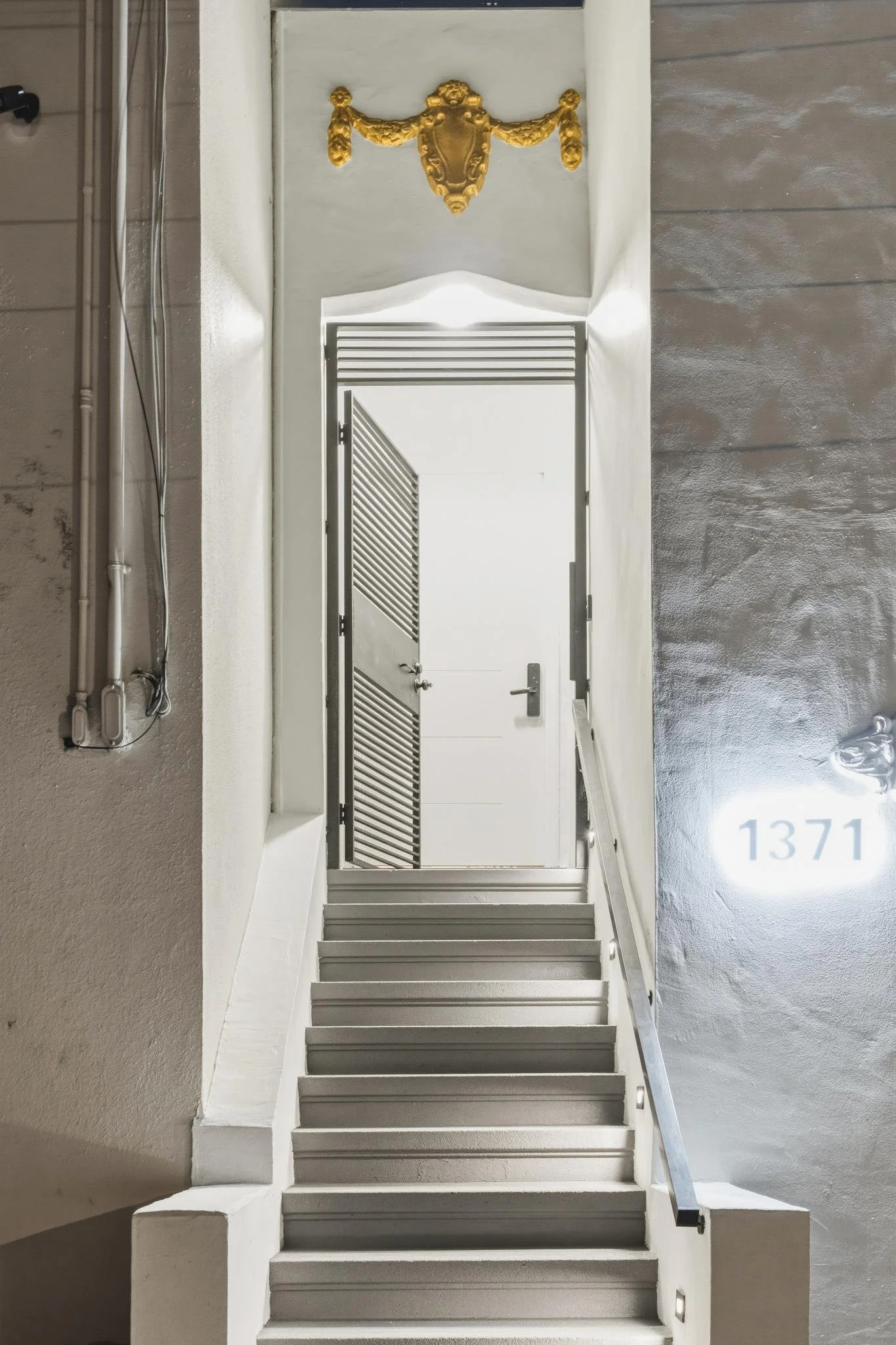 A stairwell leading up to a closed door with a security latch. The stairs are painted white, and the area is well-lit. There is a gold decorative fixture at the top of the stairway and the number 1371 illuminated on the wall to the right.