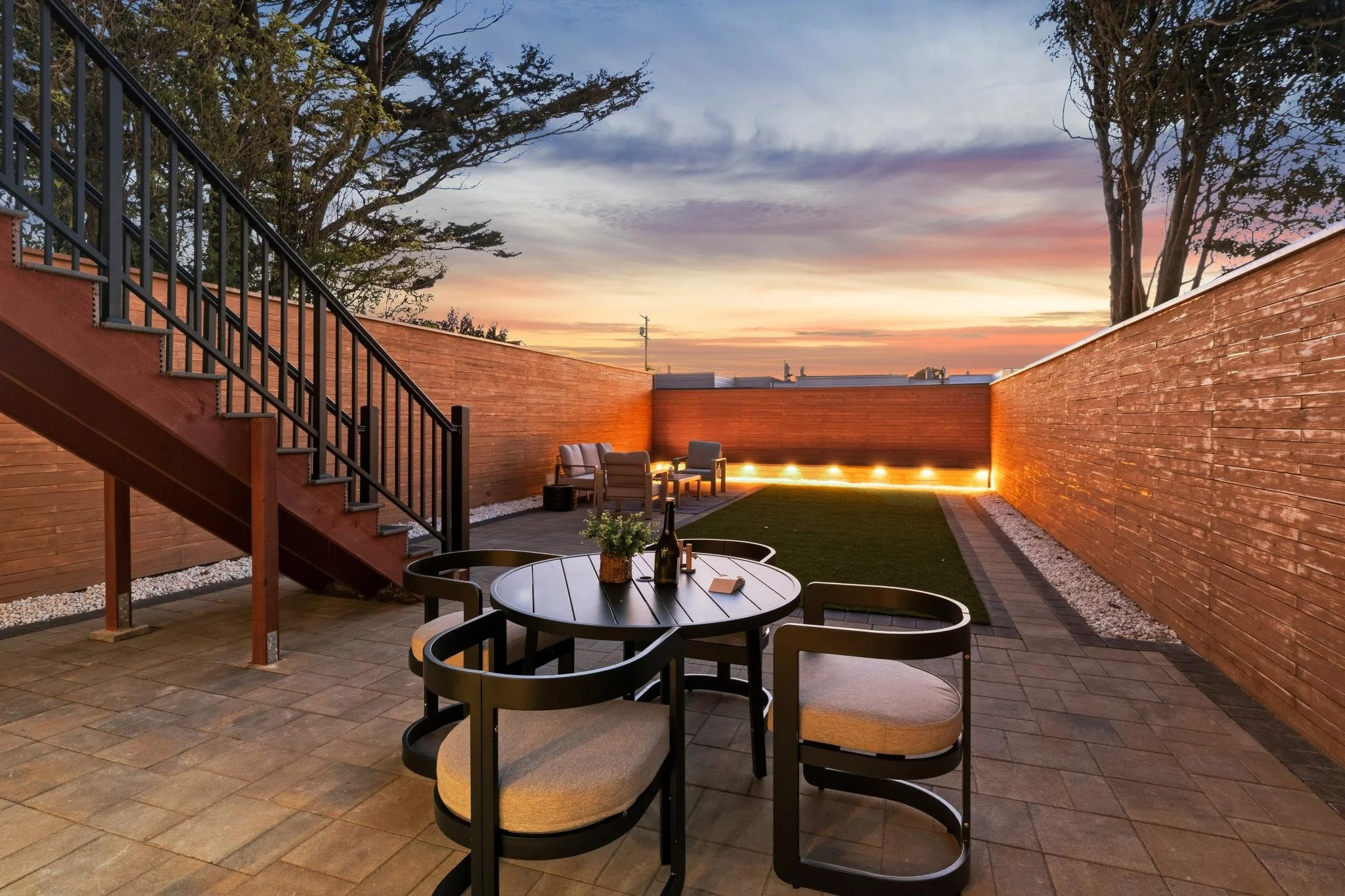 A cozy outdoor patio at sunset with a round table and four chairs, a small seating area with two chairs and a table, a grass patch, enclosed by a wooden fence with warm lighting, and a staircase leading to an upper level.