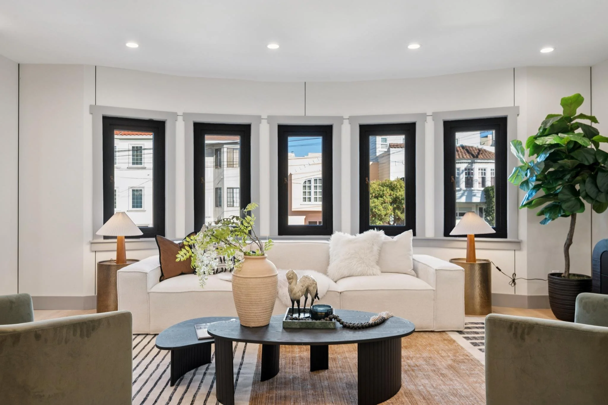 Living room with white sofa, six windows with black frames, potted plant, side lamps, coffee table with decorative items, and green chairs in the foreground.