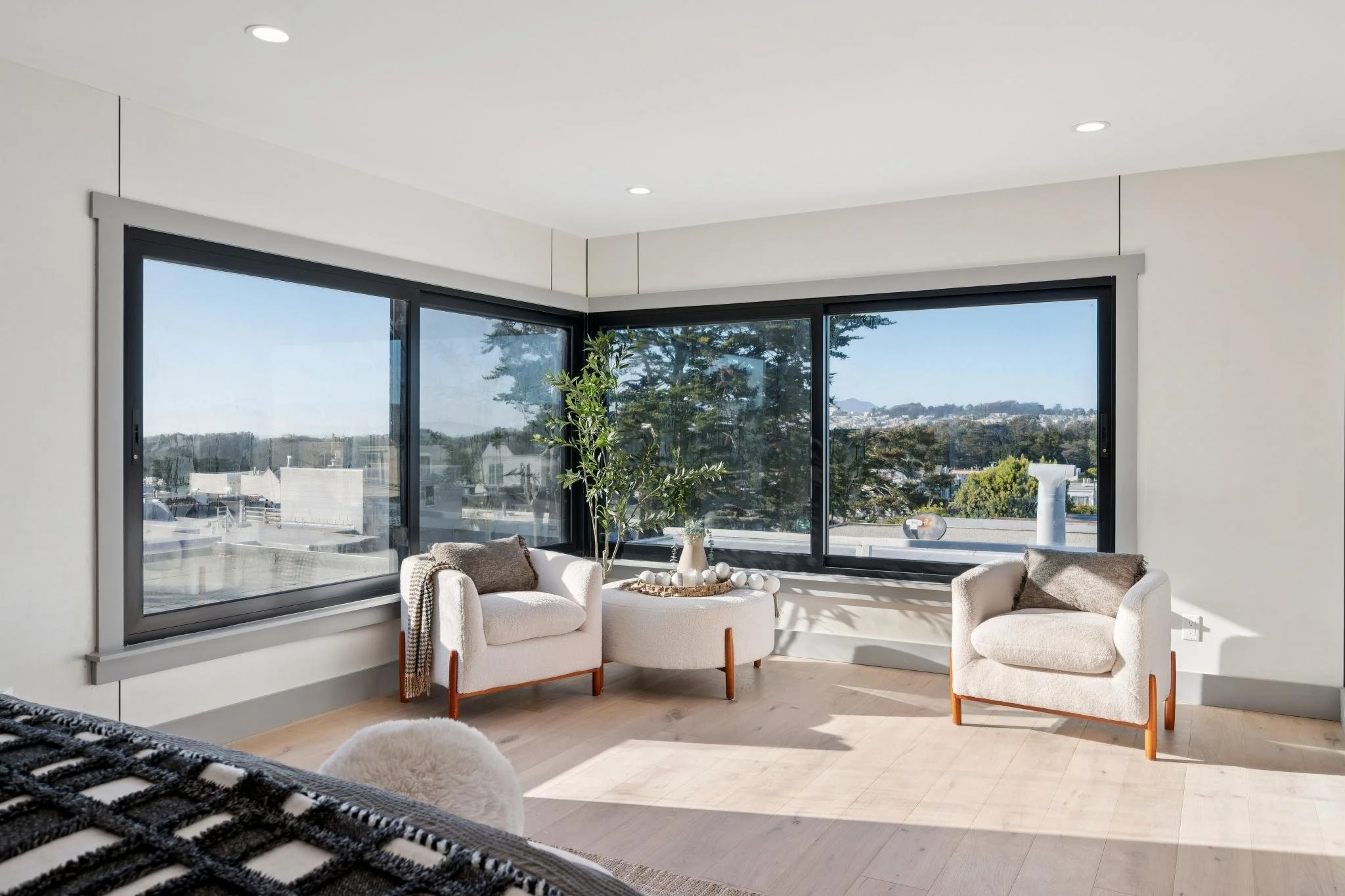 Living room with large corner windows, two cream armchairs with brown pillows, a white circular table with a plant and decorative rocks, hardwood floors, and white walls.