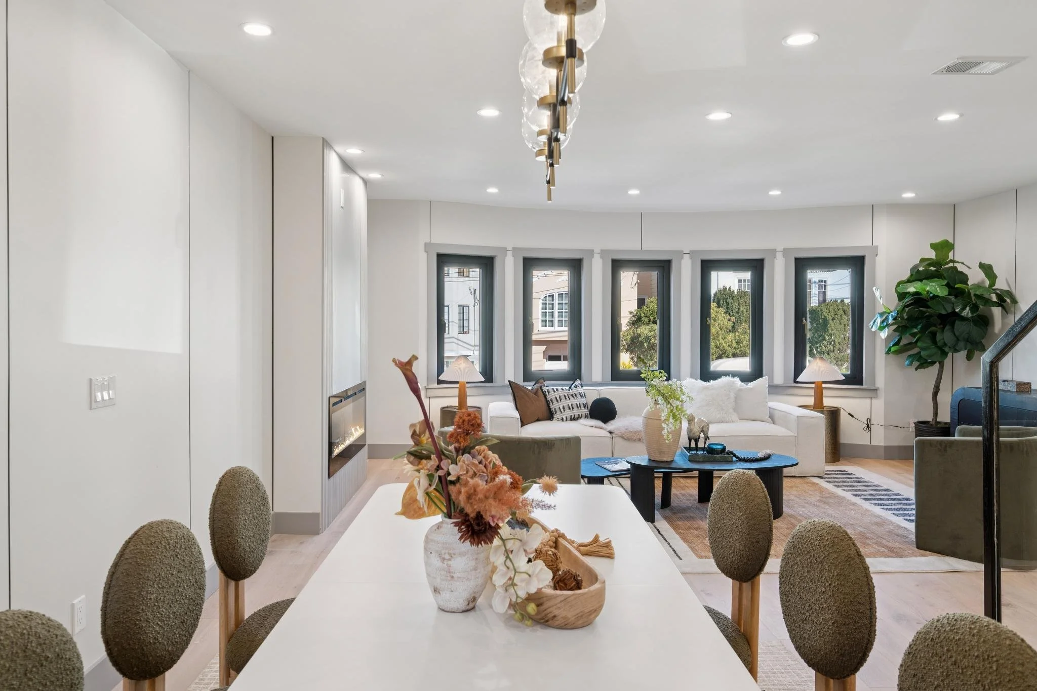 Living room and dining area with white walls, a rectangular white table with six chairs, a beige vase with flowers on the table, a white sofa with pillows, a coffee table with decorative items, and large windows showing trees outside.
