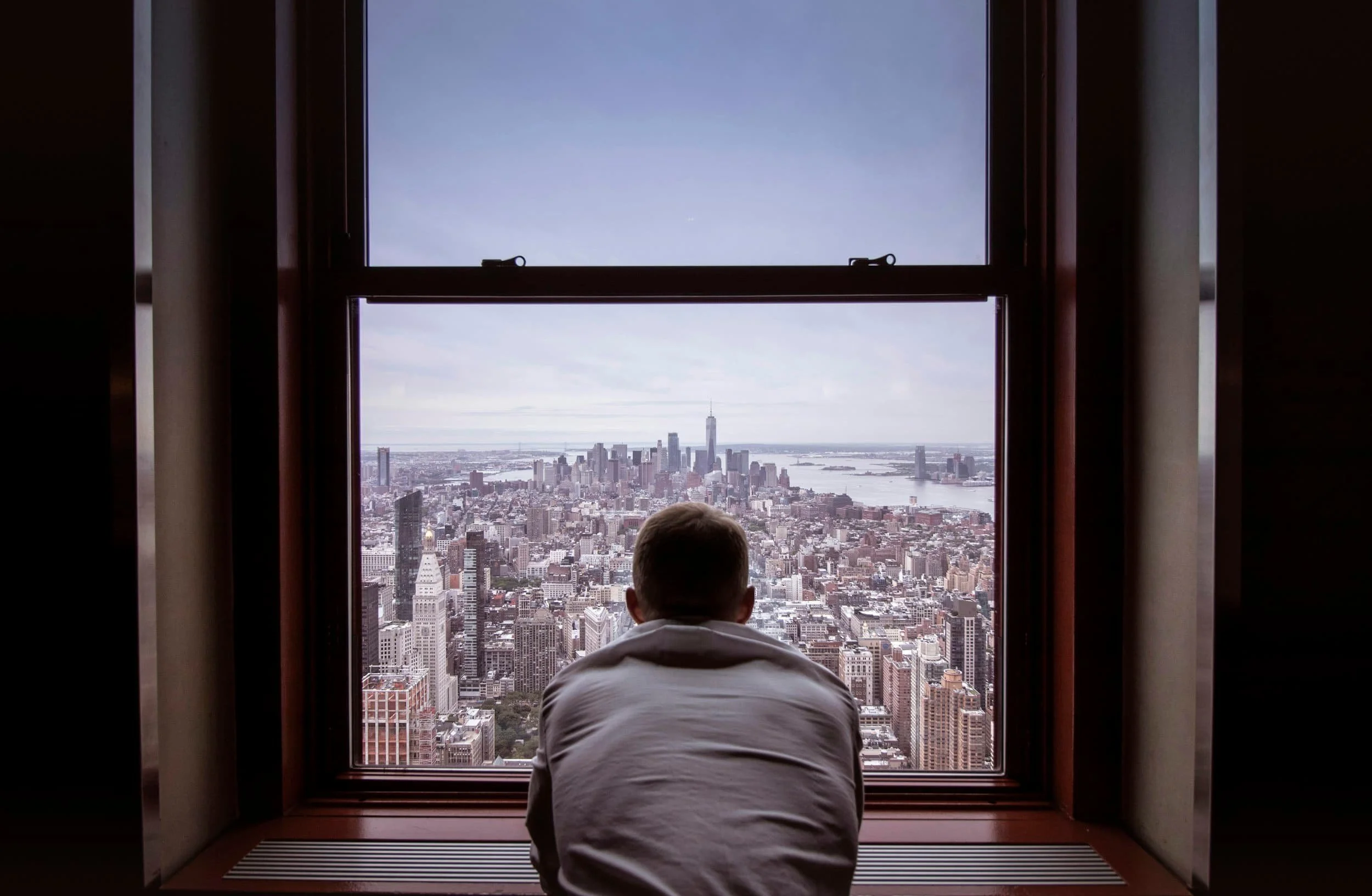 A boy in a white jacket sitting on a windowsill, looking out at the New York City skyline including the One World Trade Center and the Hudson River.