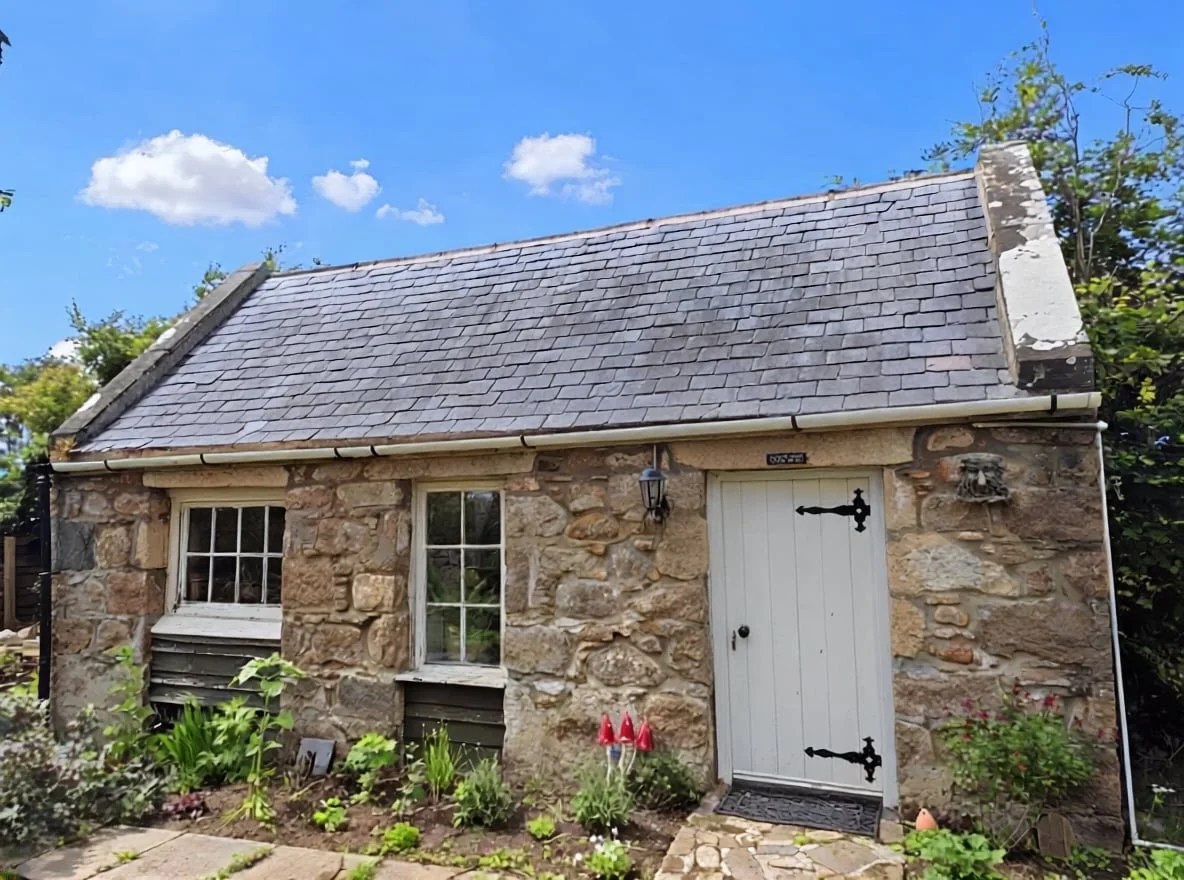 A small stone cottage with a slate roof, white wooden door, and two small windows, surrounded by a garden with flowers and plants, under a bright blue sky with a few clouds.
