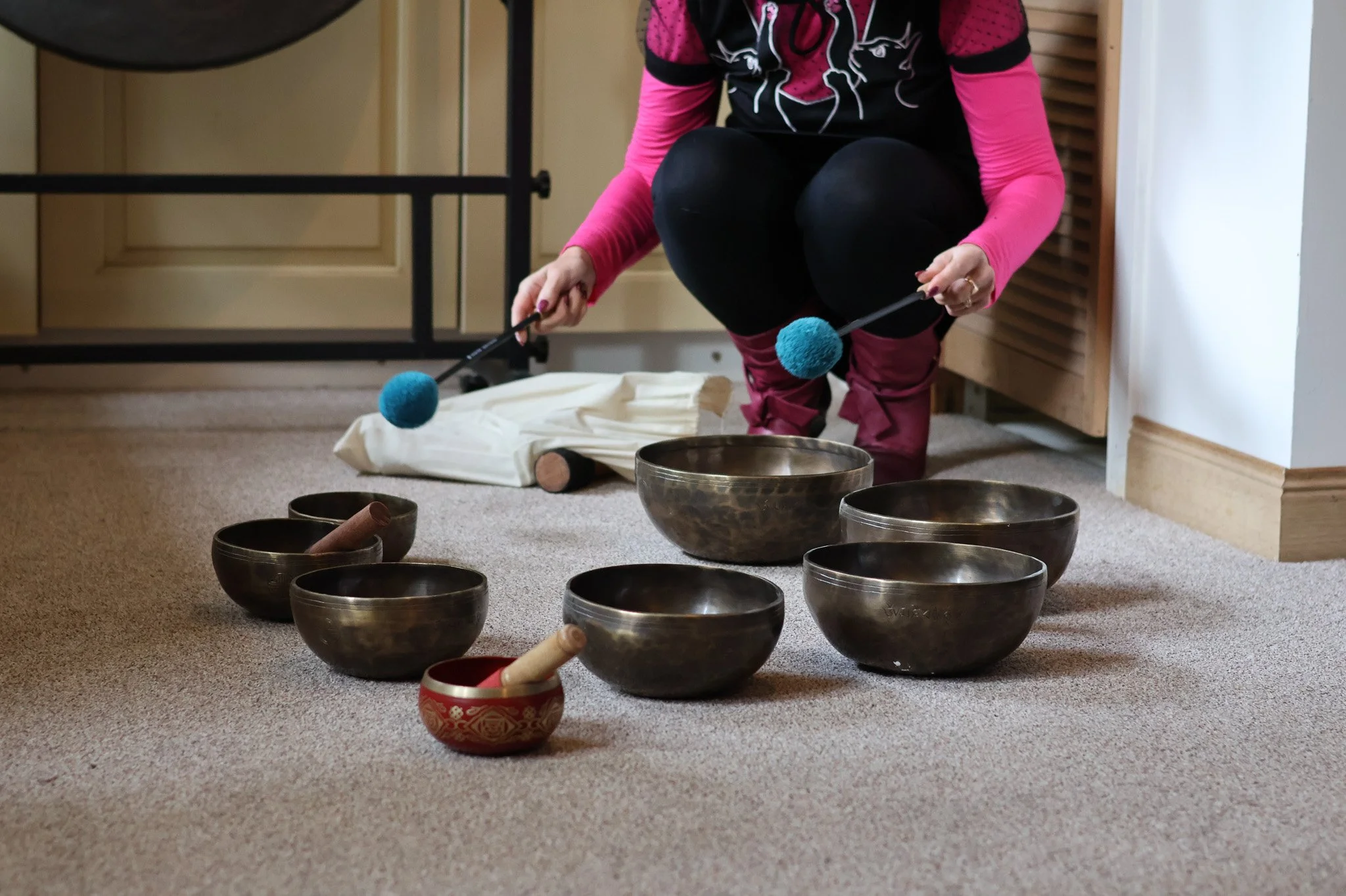 Person crouching and holding a mallet, surrounded by singing bowls on the floor, likely preparing for a sound therapy or meditation session.