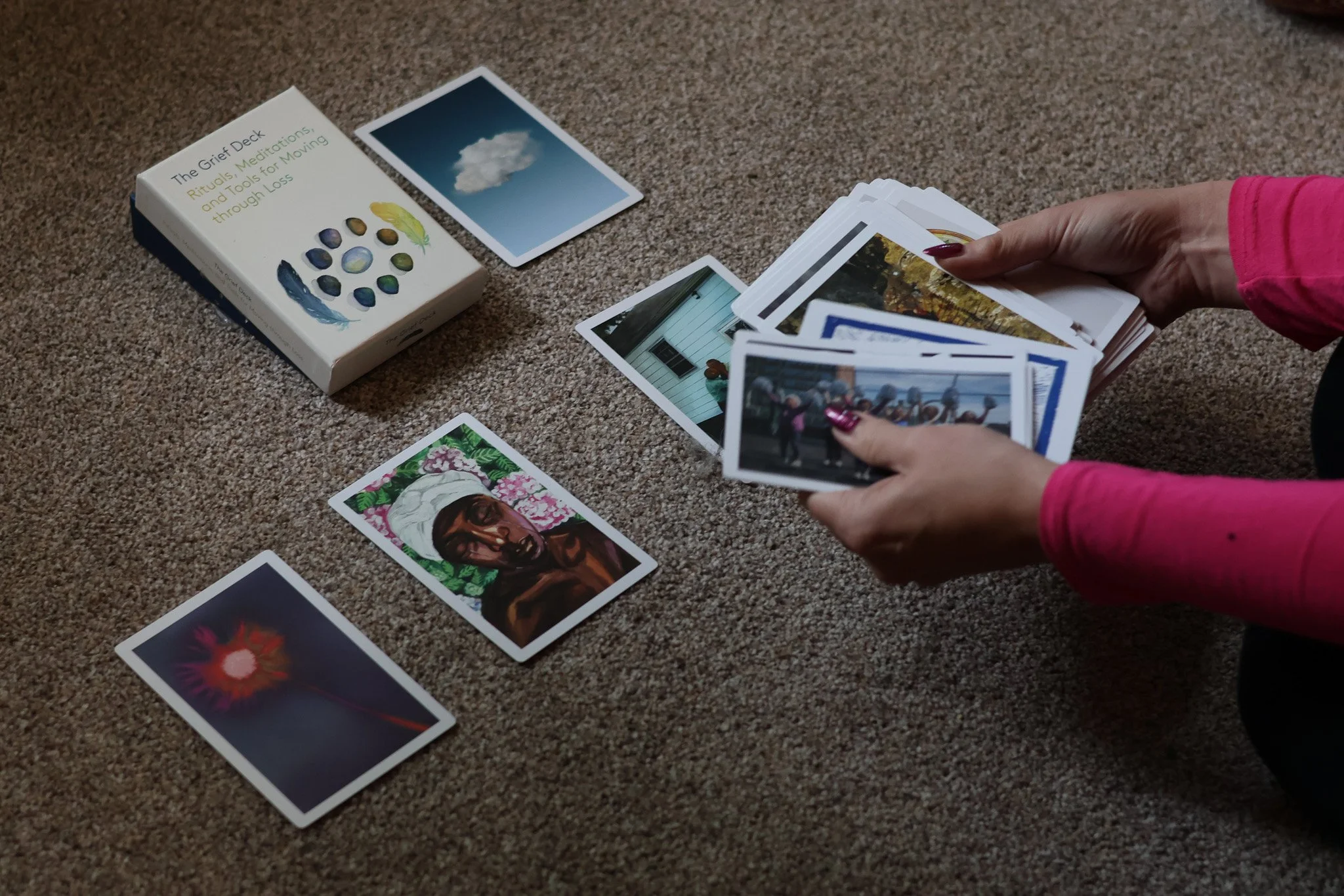 A person wearing a pink long-sleeve shirt holding a stack of photographs over a carpeted floor, with several other photographs laid out. There is a deck of cards titled 'The Grief Deck' with the subtitle 'Rituals, Meditations, and tools for Moving through Loss' and a card with an image of a cloud on it.