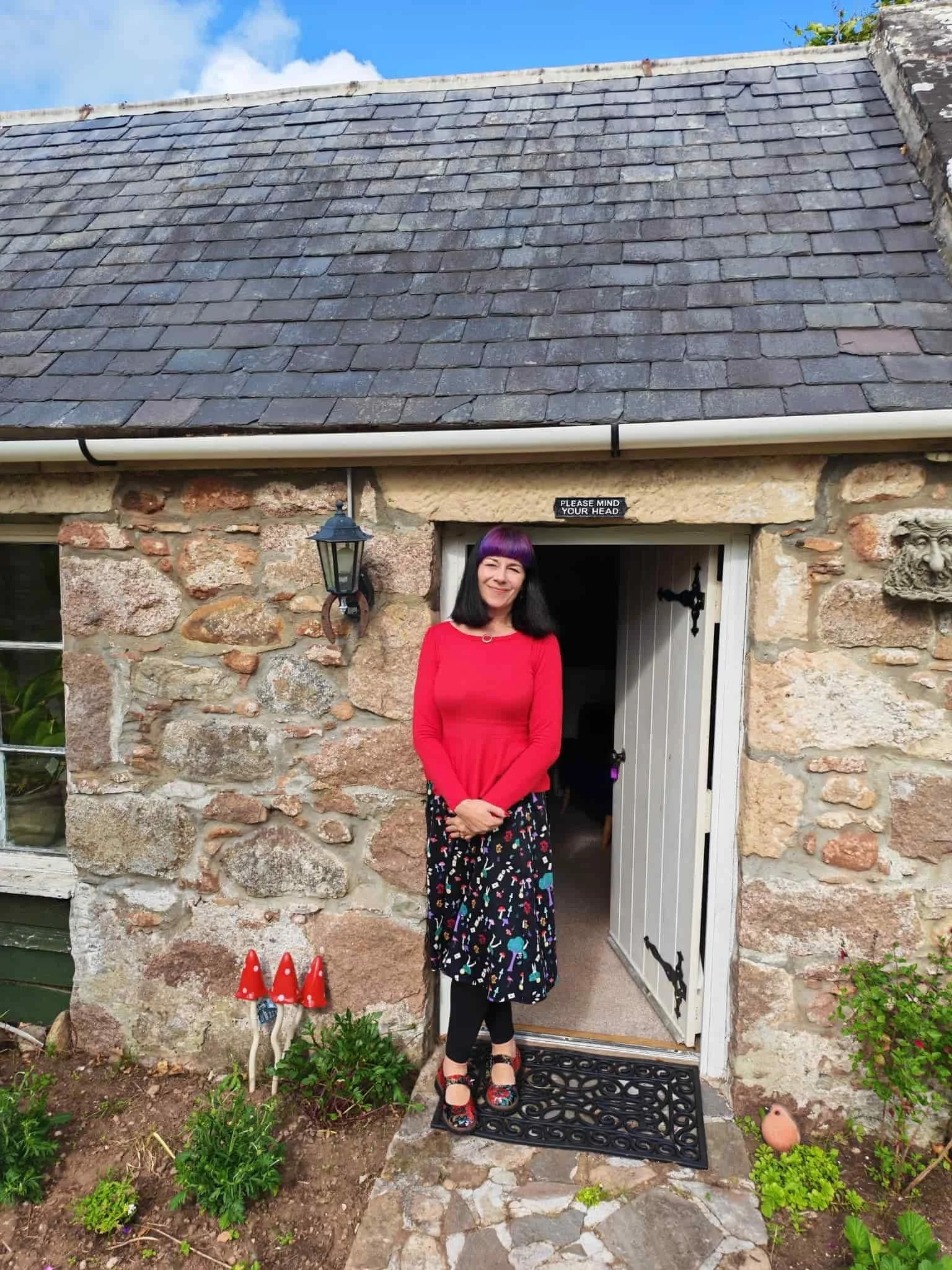 A woman with black hair and purple bangs wearing a red top and a long skirt with colorful pattern, standing in front of a stone house with a white door, black lantern, garden plants, and a small decorative stone sculpture outside.