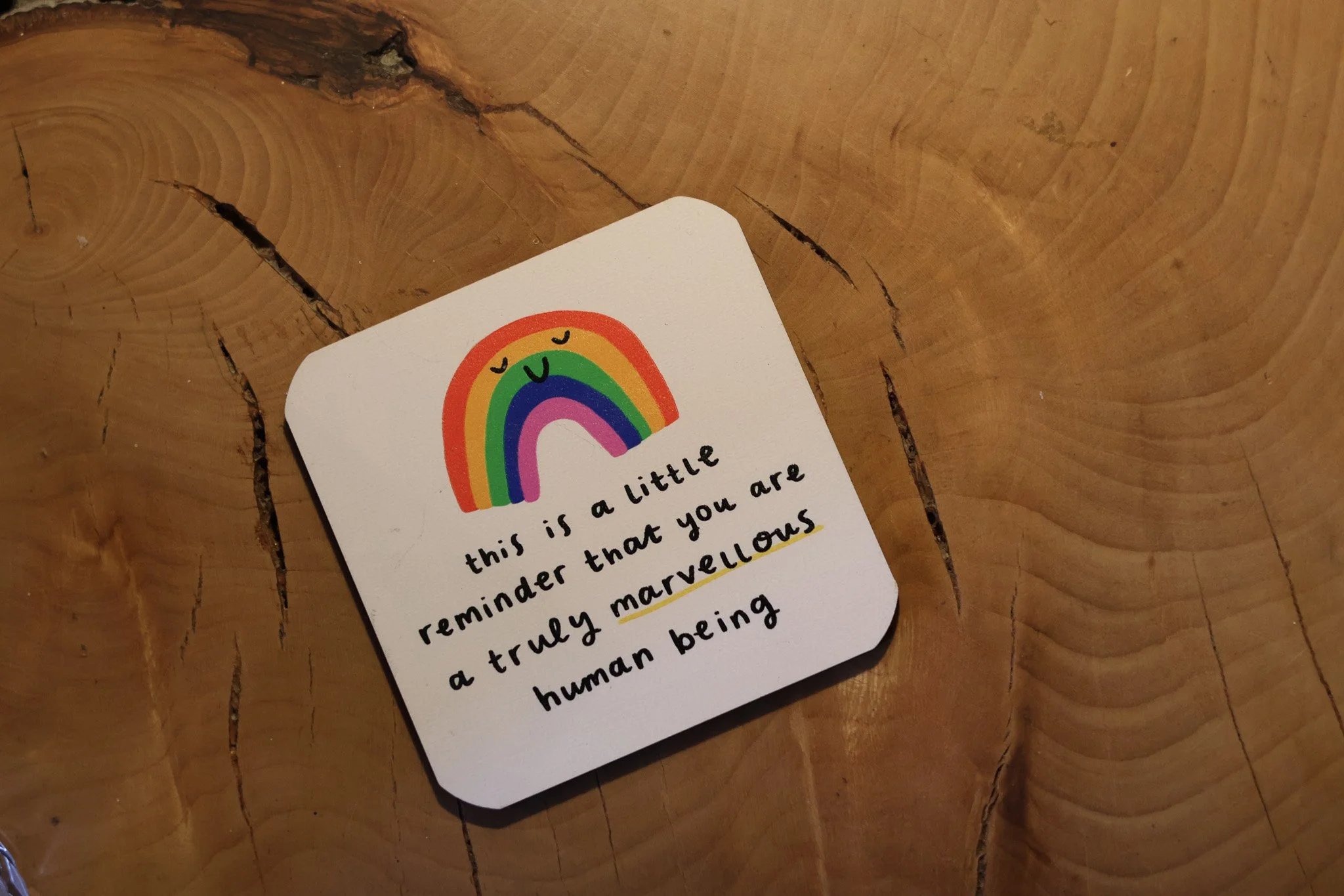 Close-up photo of a colorful rainbow sticker with a smiling face, placed on a wooden surface. Part of a handwritten note with black ink is visible underneath the sticker, with some words highlighted in yellow.