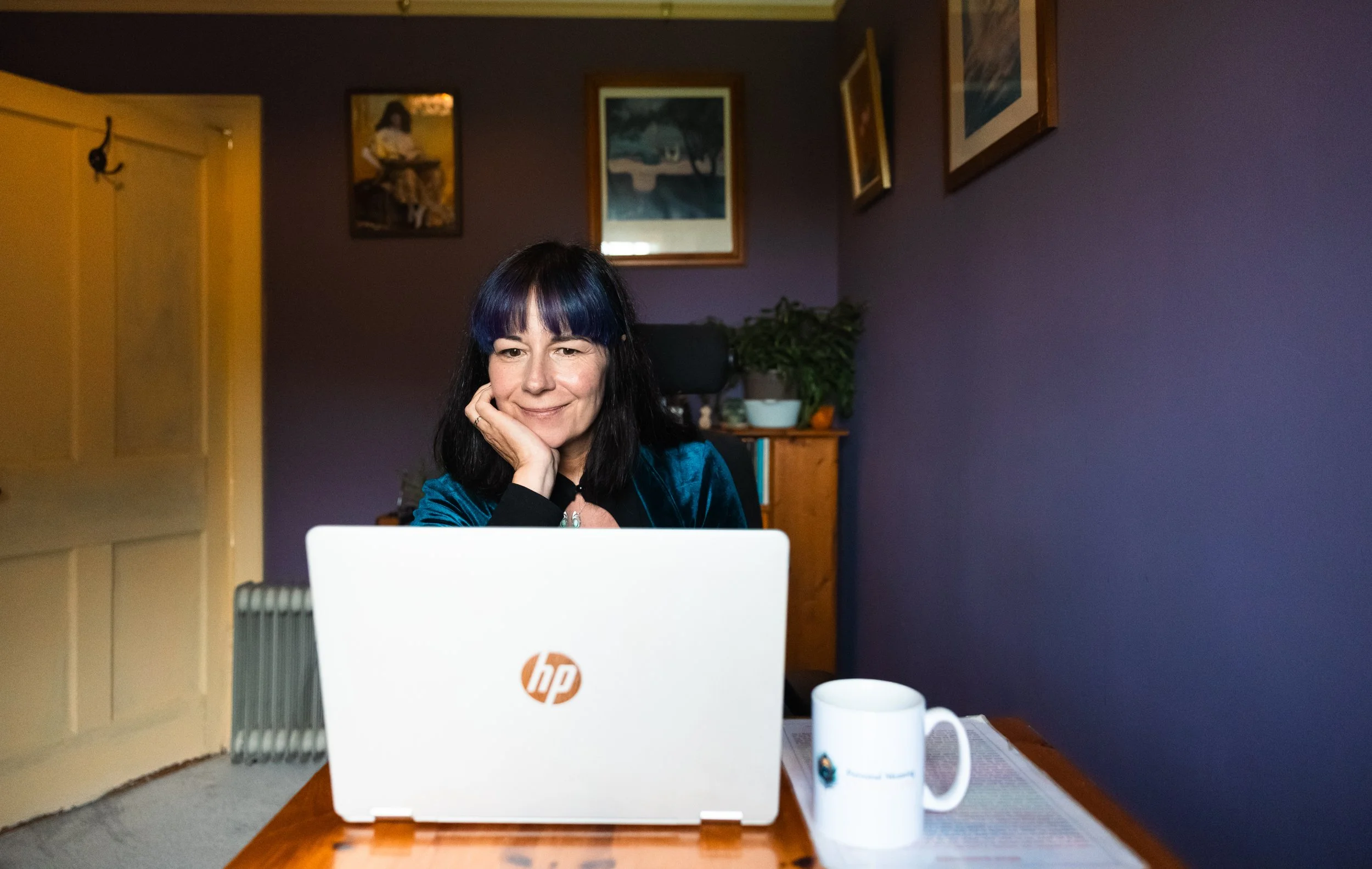 A woman with black hair and bangs sitting at a desk, resting her chin on her hand, looking at a white HP laptop. The background features a purple wall, framed artwork, and houseplants. A coffee mug and papers are on the desk.
