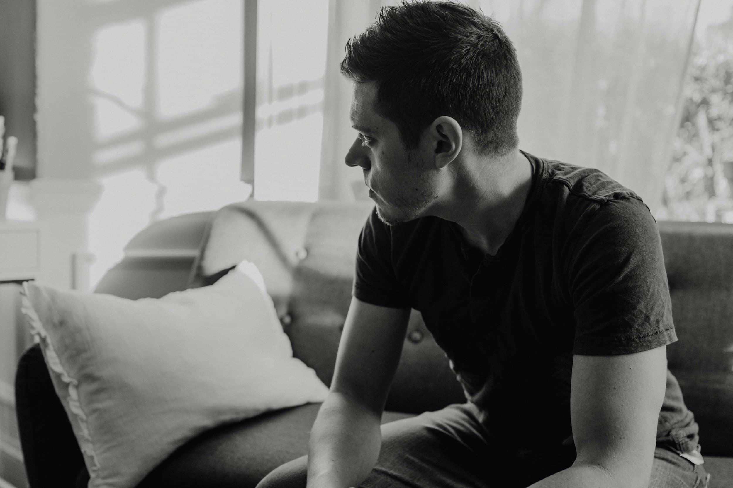 A young man with dark hair and a beard sitting on a bed in a sunlit room, looking down and leaning forward with his elbows resting on his knees. The room has a window with sunlight streaming in, casting shadows on the wall.