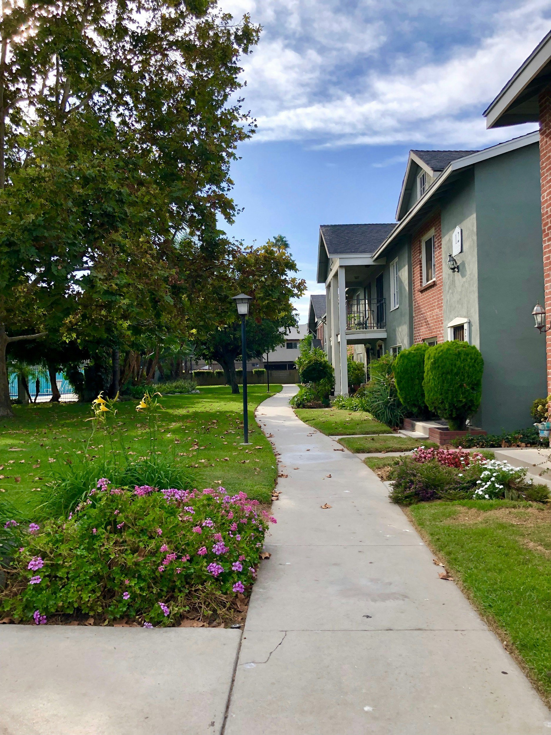 A sidewalk winds through a residential neighborhood with well-maintained gardens and vibrant flowers on both sides, flanked by modern houses with lush green lawns, trees, and shrubs, under a partly cloudy sky.