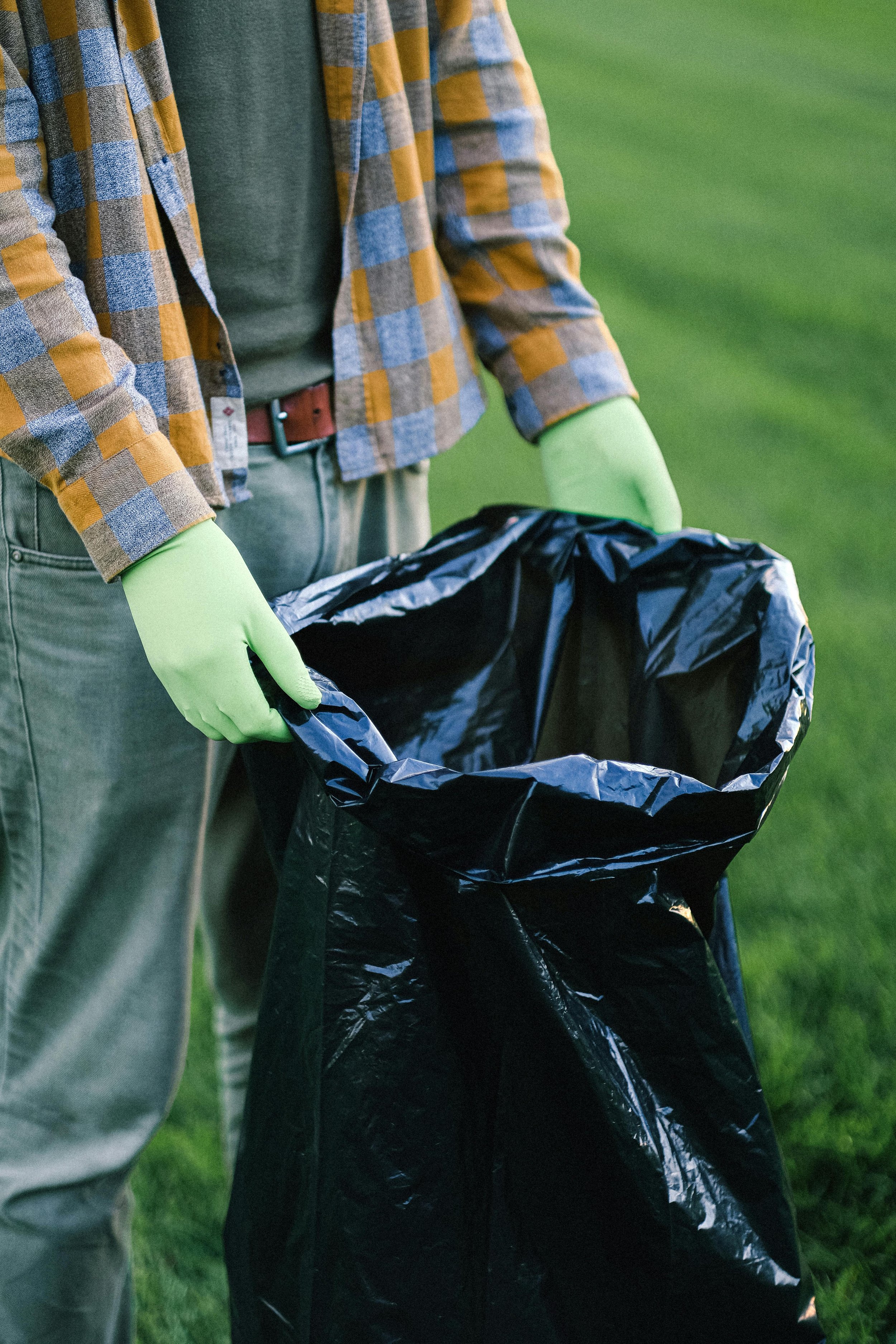 Person wearing gloves and a plaid shirt holding a black trash bag outdoors on green grass.