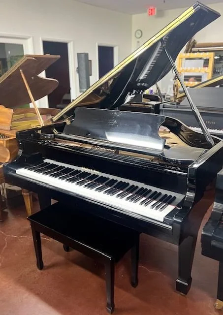 A black grand piano with its lid open on a wooden floor in a music studio or showroom, surrounded by other pianos and musical equipment.