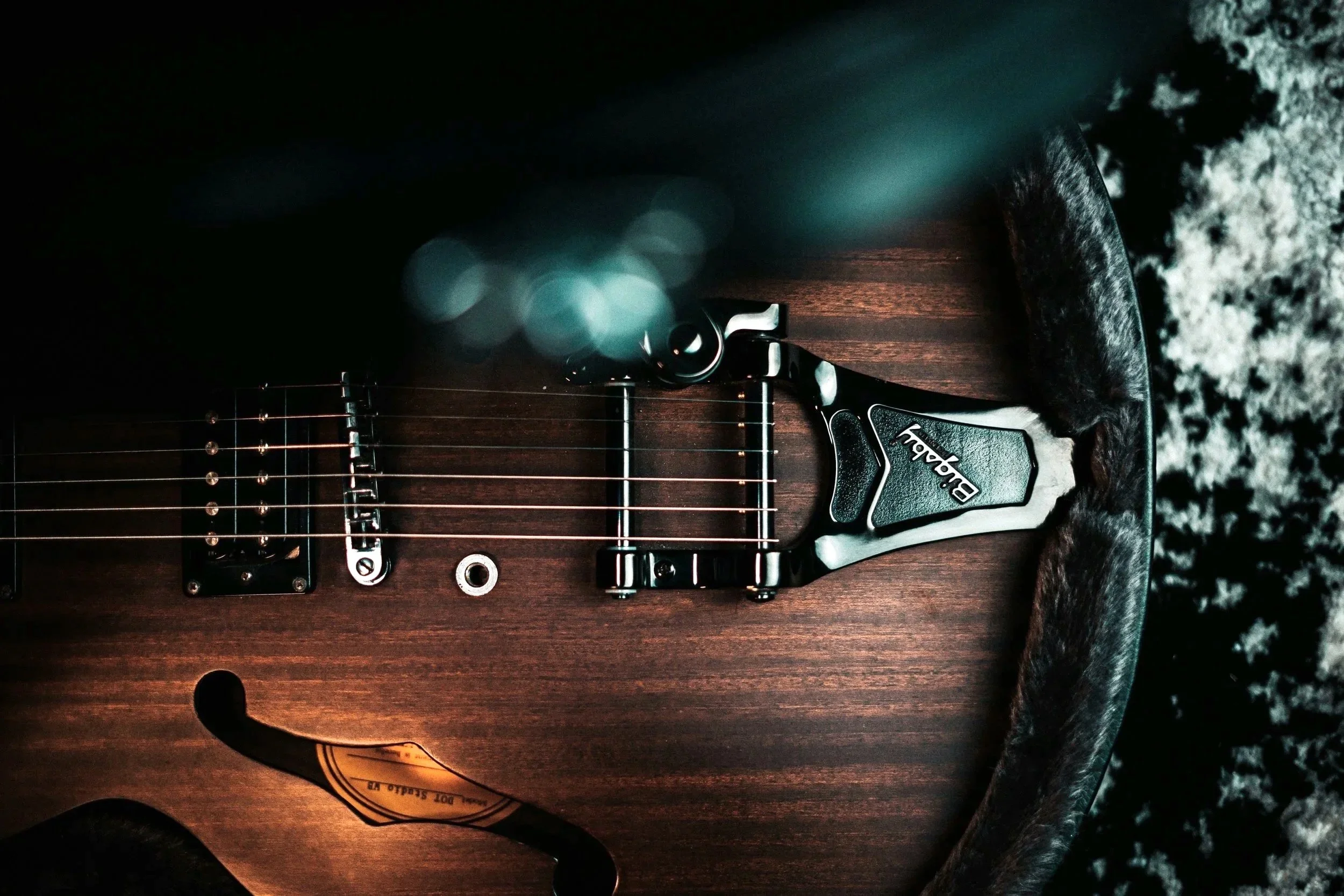 Close-up of an electric guitar's body, showing strings, pickups, and a control knob, resting on a dark surface with a textured black and white background.